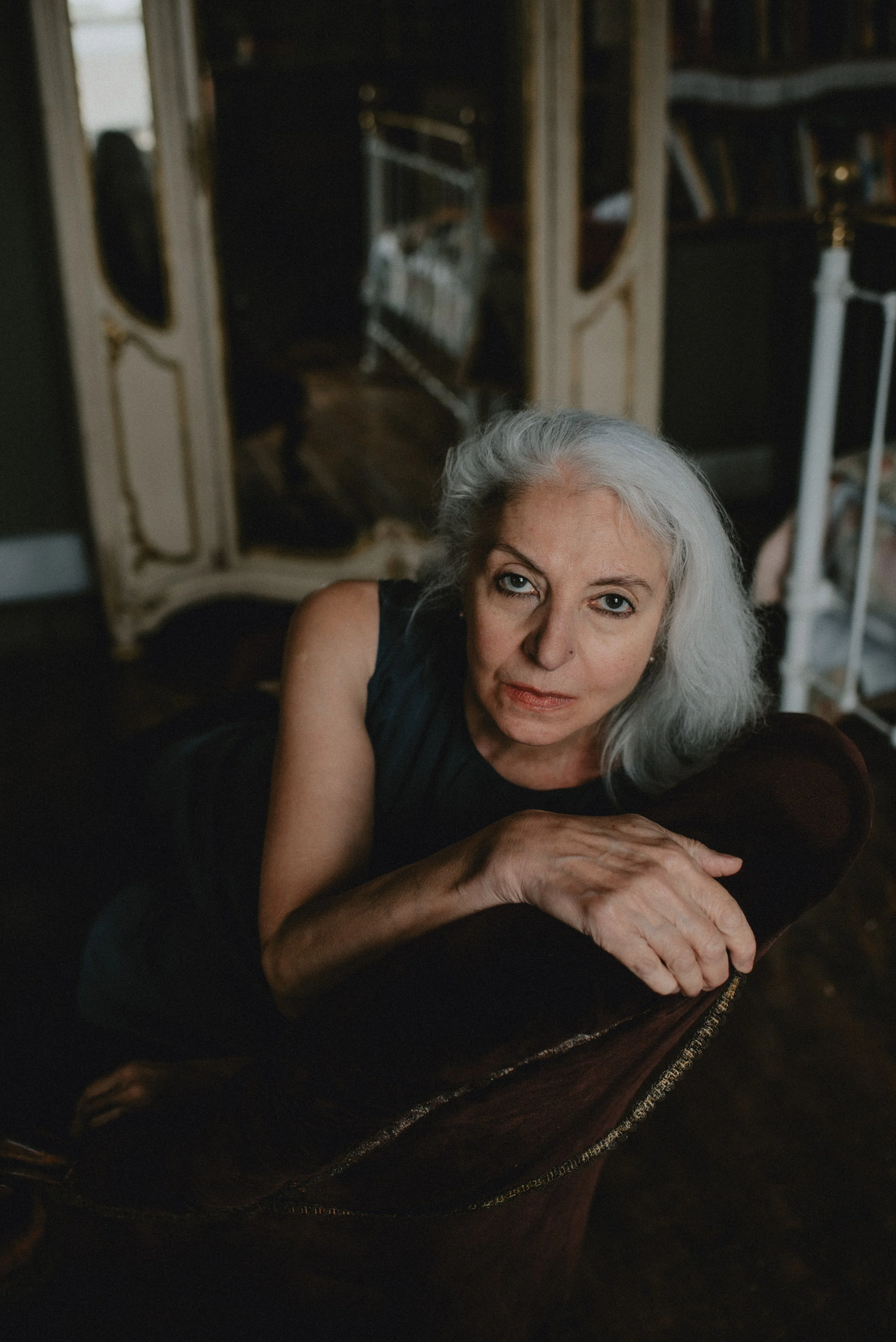 A woman with gray hair lying on a dark velvet armchair, looking directly at the camera, in a room with bookshelves and an ornate mirror in the background.