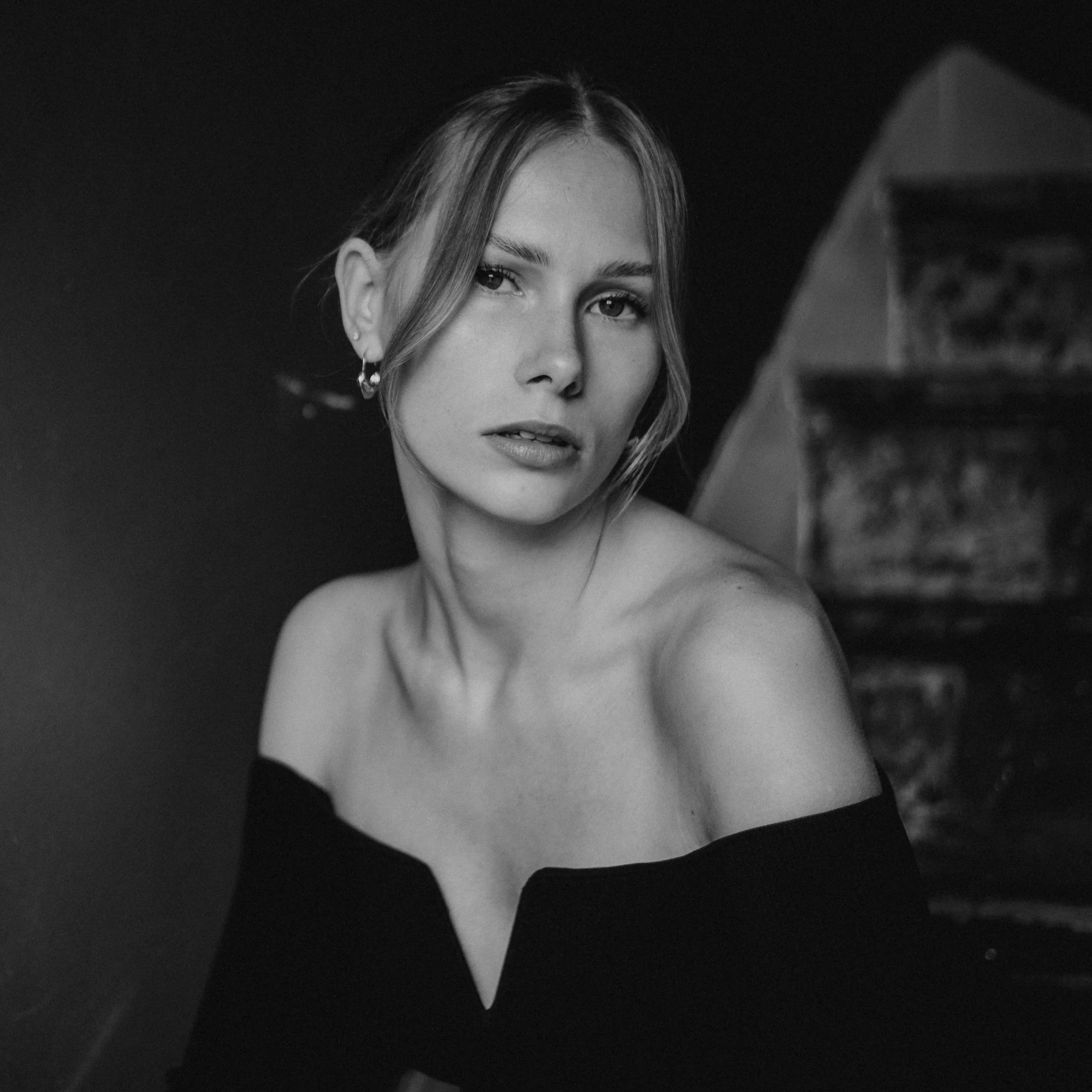 Black and white portrait of a woman with shoulder-length hair, wearing an off-the-shoulder top, looking at the camera with a serious expression, in front of a dark background with a blurred staircase.