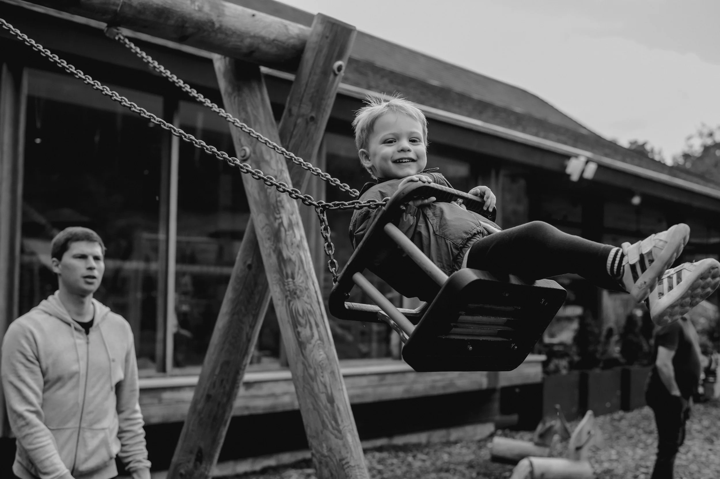 A young child smiling and swinging on a playground swing, with an adult watching nearby. The scene is outdoors with a wooden building in the background.