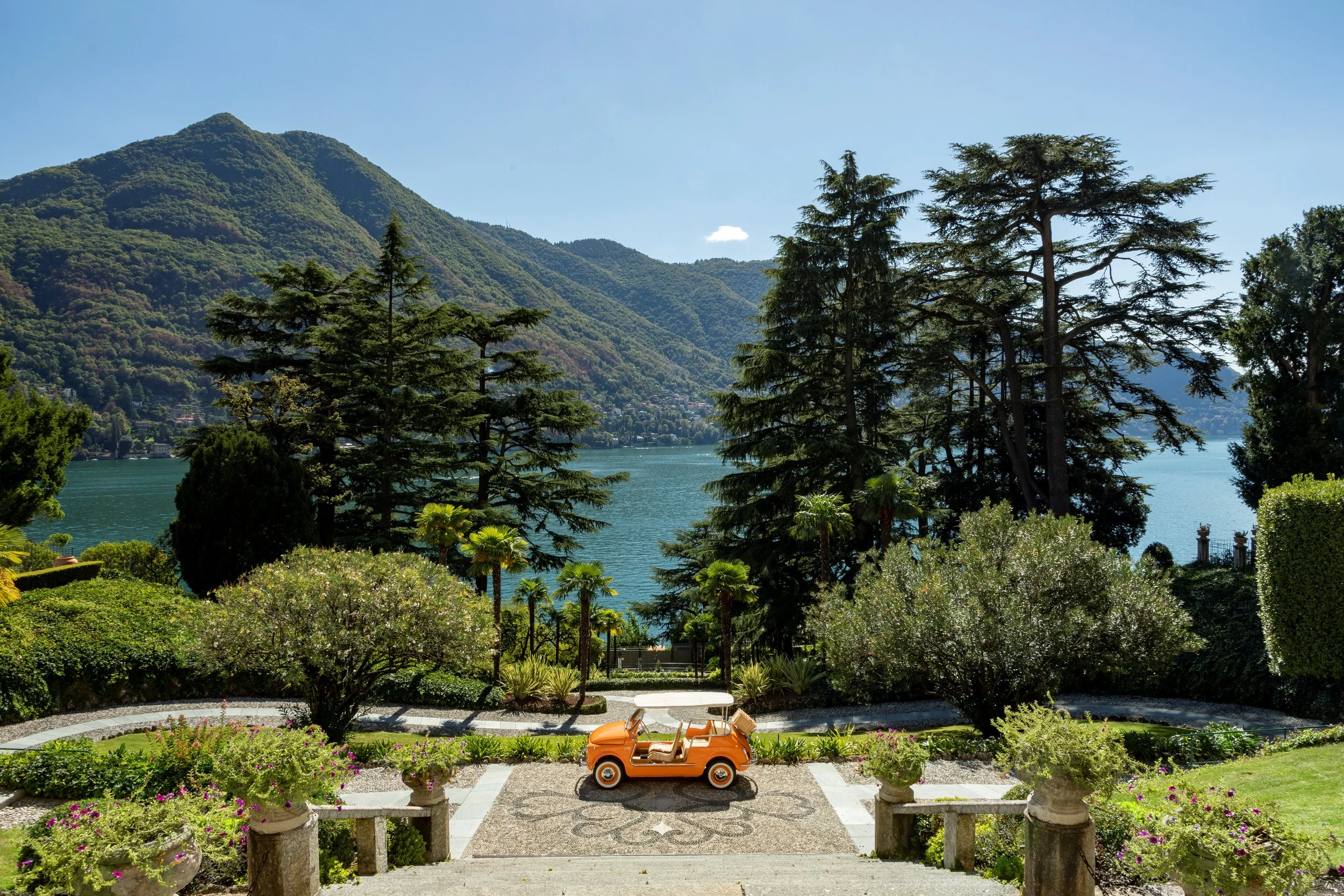 A scenic view of a lakeside mountain landscape framed by lush green trees and shrubs, with a small, vintage orange toy car placed on a decorative stone and tile patio in the foreground.
