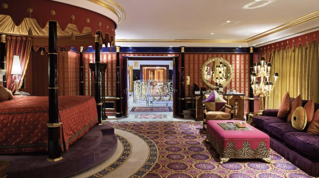 Luxurious hotel bedroom suite with a four-poster bed, ornate curtains, a purple velvet sofa, a pink upholstered ottoman, and gold accents.