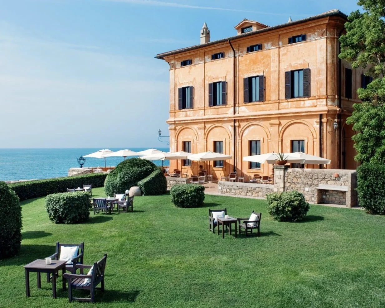 A large orange building with black shutters on the windows, overlooking the ocean. The foreground shows a well-manicured lawn with outdoor furniture and white umbrellas, suggesting a seaside resort or hotel.