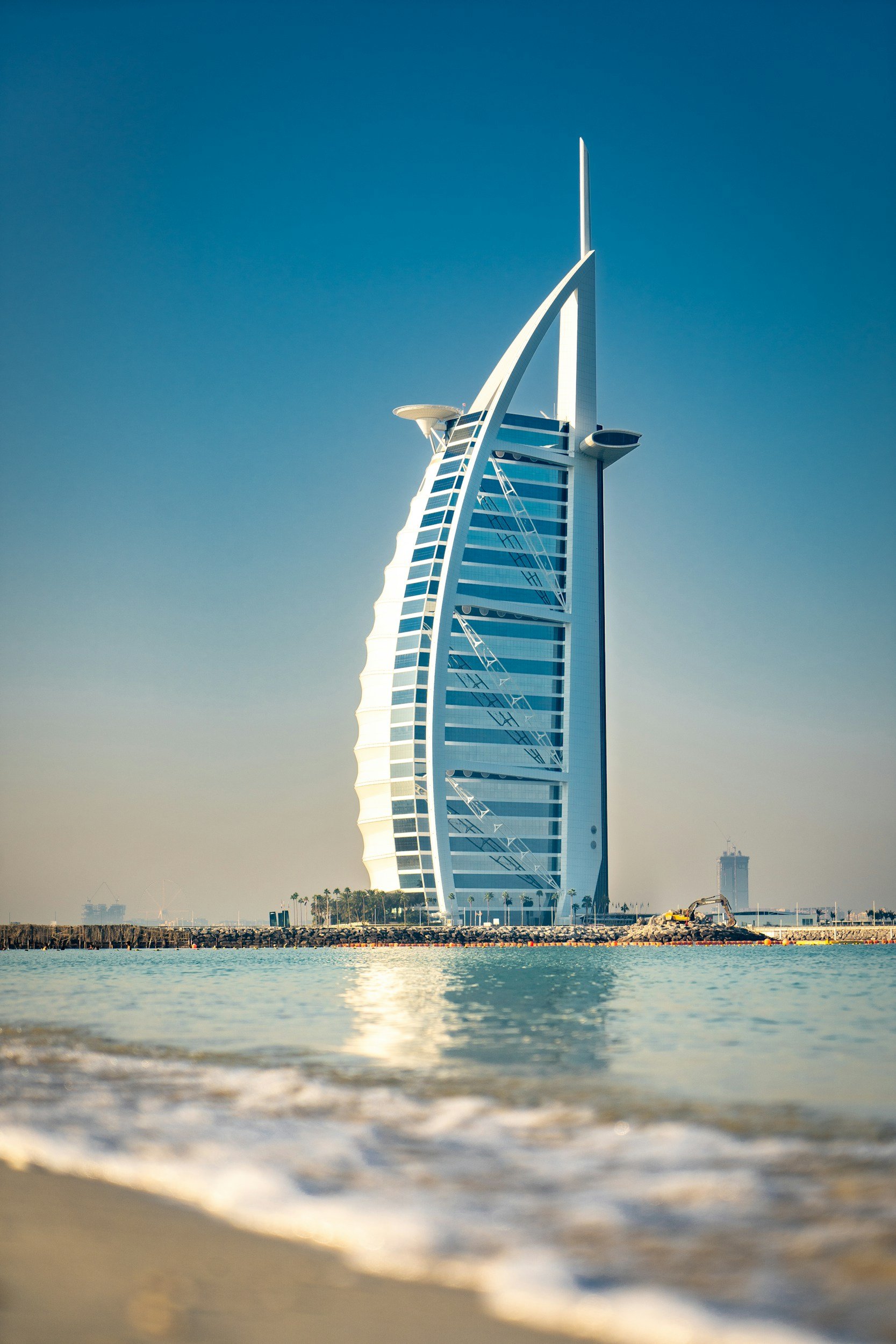 The image shows the Burj Al Arab hotel in Dubai, a distinctive sail-shaped luxury hotel located by the water, with a sandy beach and calm sea in the foreground.