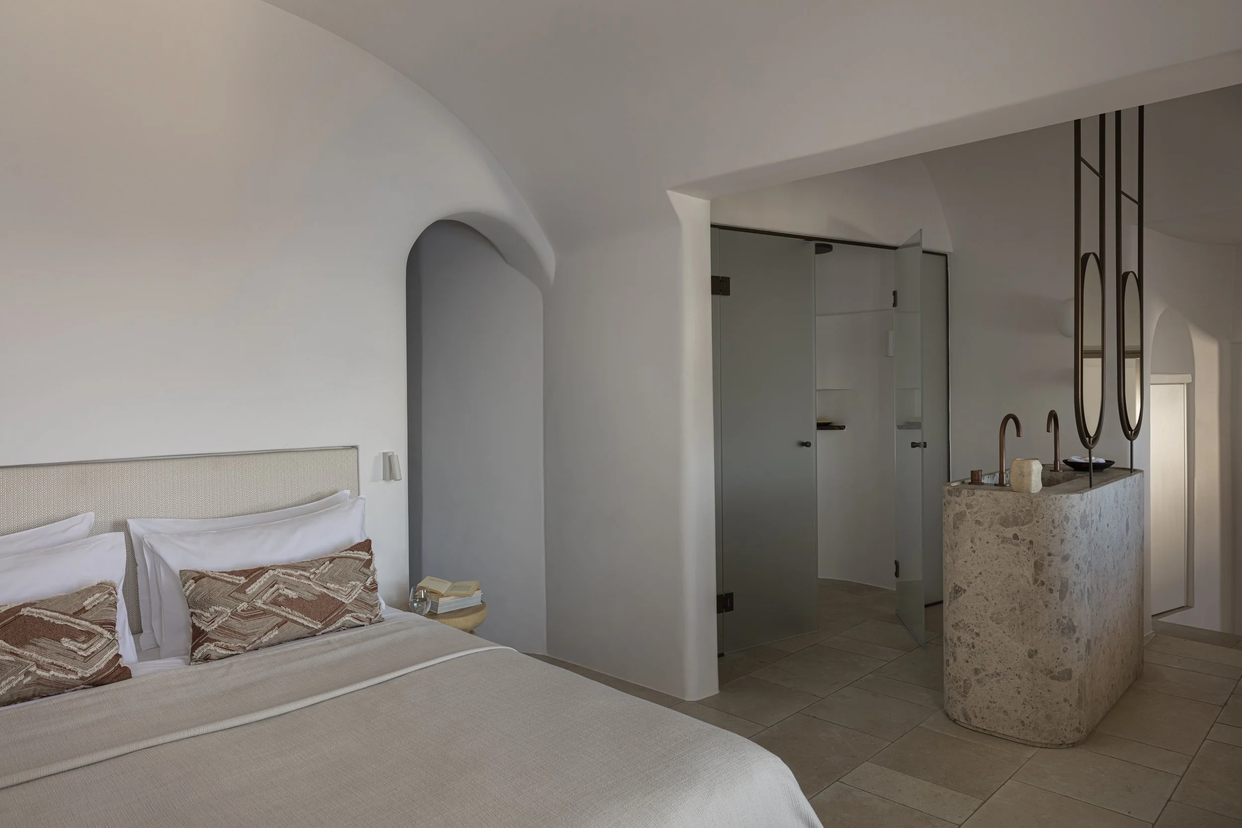 A minimalist hotel bedroom with a bed adorned with decorative pillows, a small bedside table, and a built-in nook. The room features white smooth walls, arched doorways, and a textured stone bar with two sinks, copper faucets, and modern pendant ligh