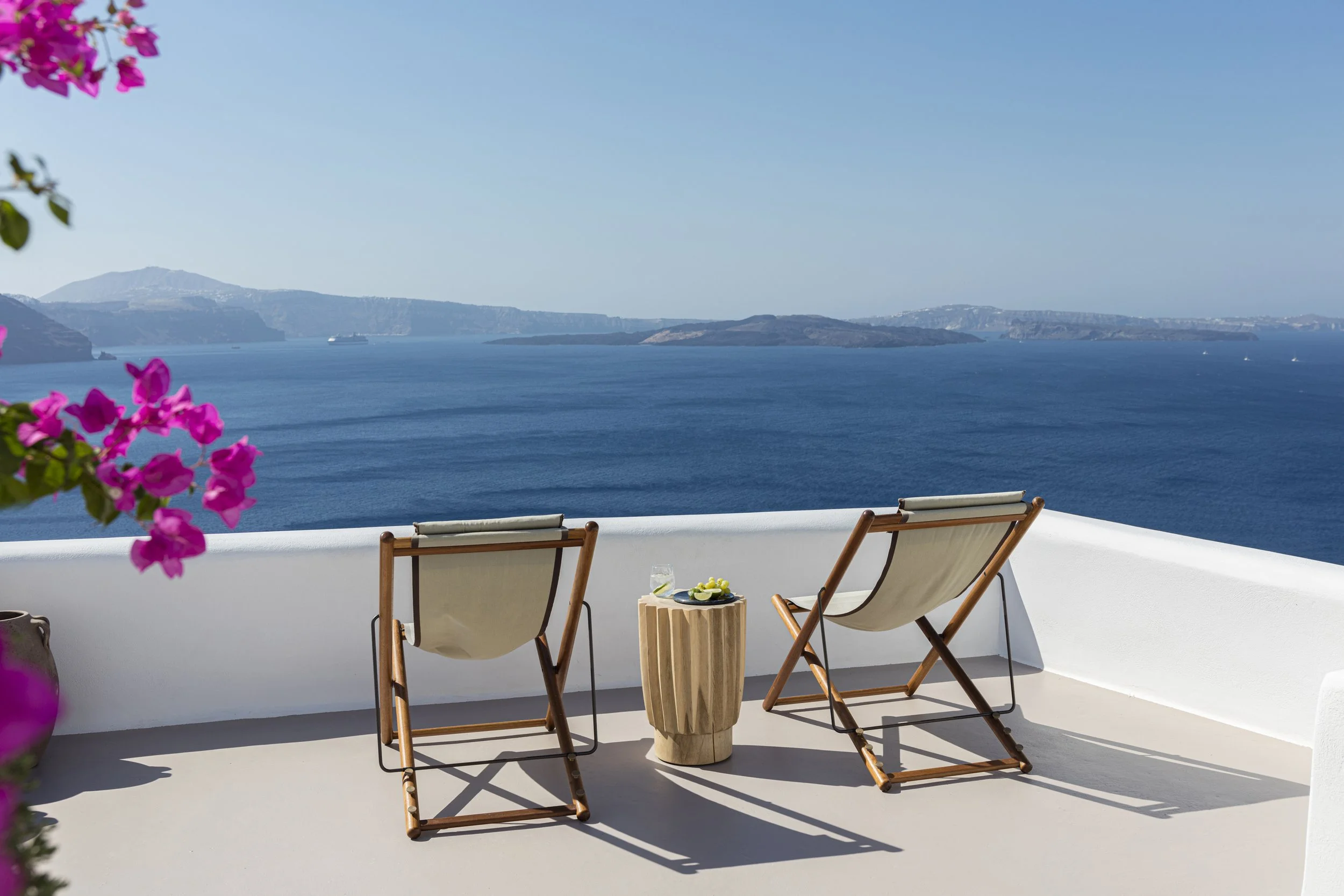 Two wooden lounge chairs with beige fabric seats facing a blue sea and distant islands, with a small wooden table between them holding a glass of water, on a white patio with a white wall and pink flowers on the left.