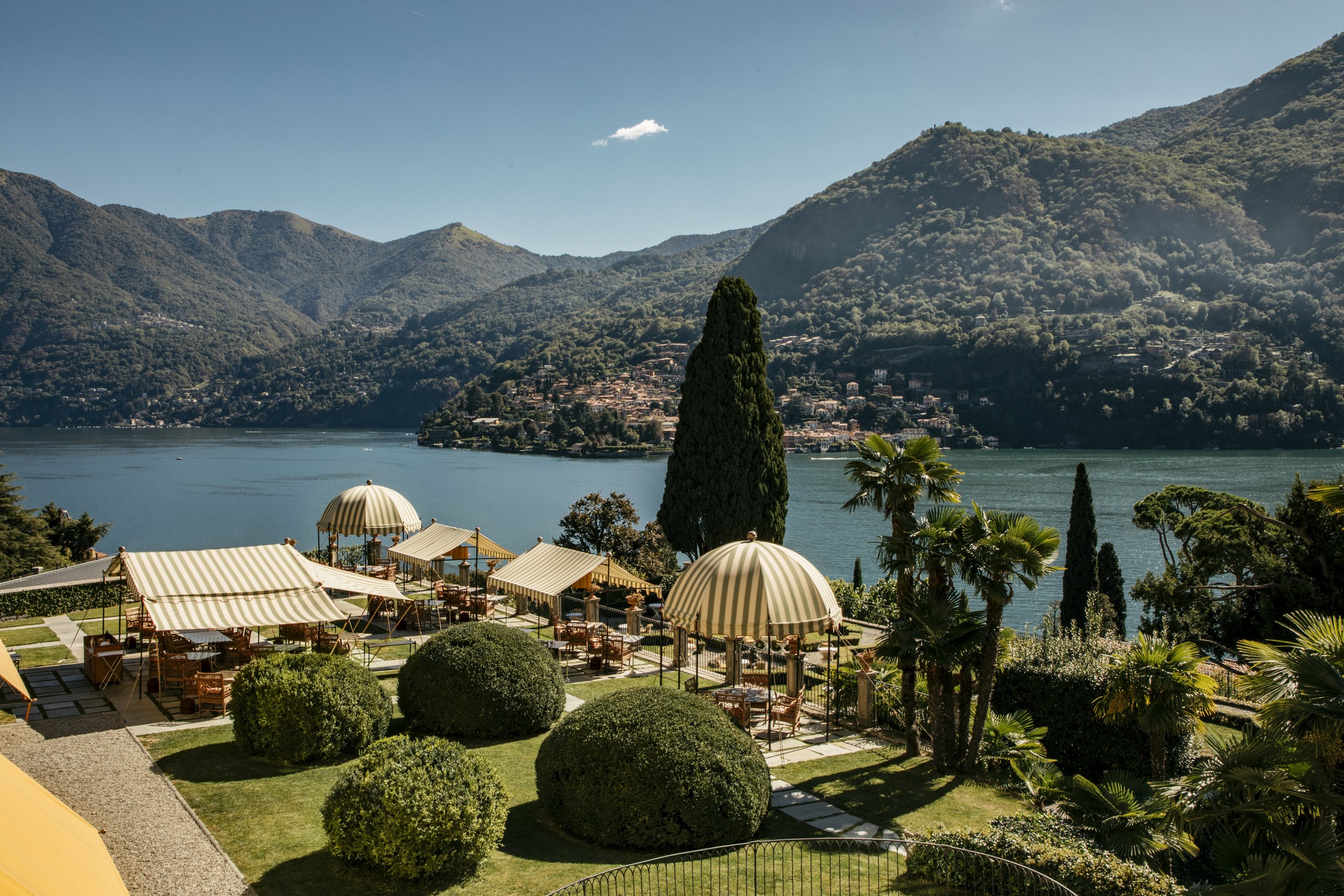 Outdoor patio with striped umbrellas and seating overlooking a lake surrounded by green mountains.