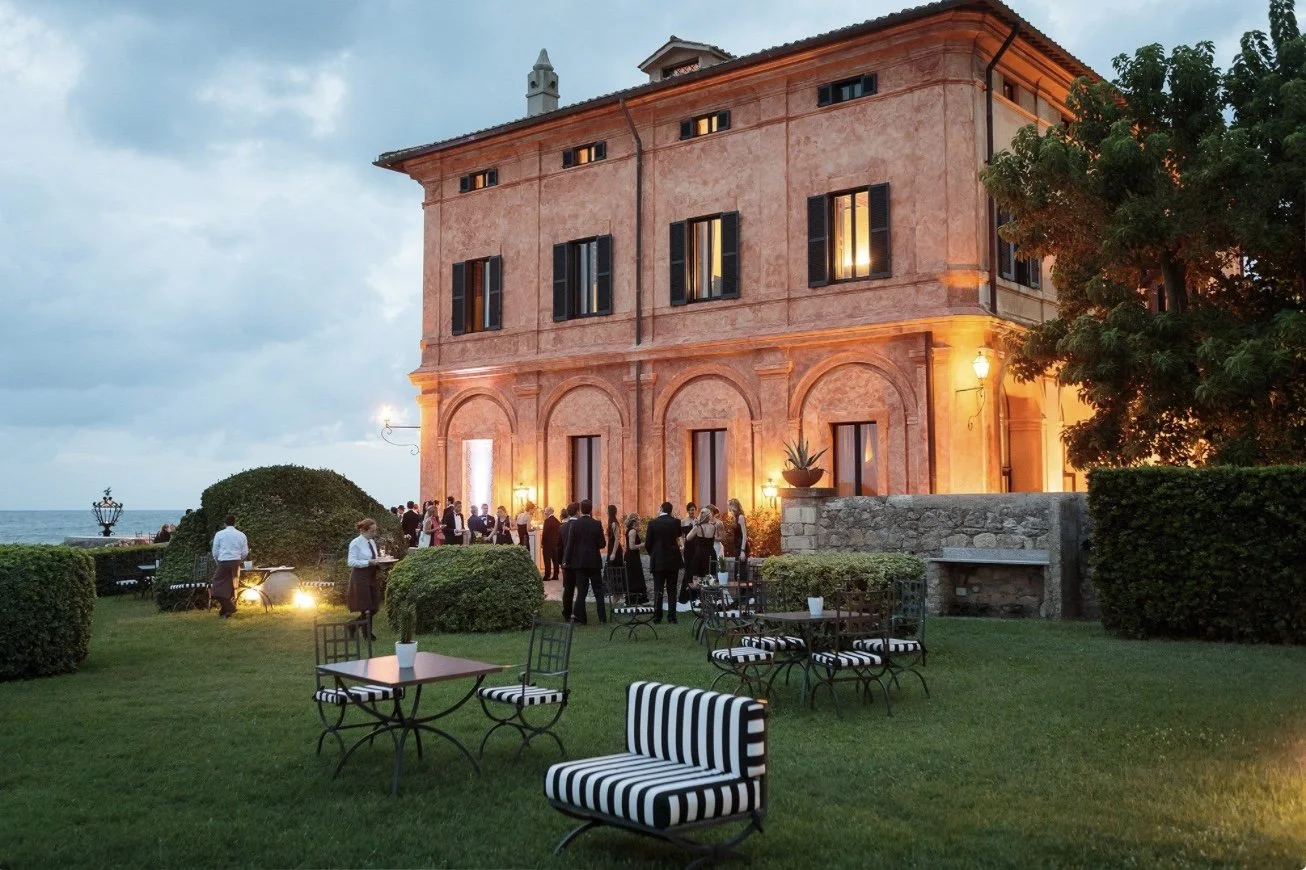 An outdoor evening event gathering with people in formal attire on a lawn in front of a large, illuminated historic building with shutters and a stone wall, overlooking the sea.