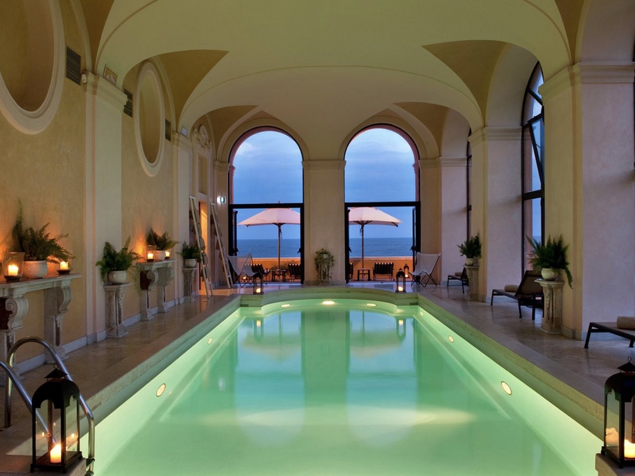 Indoor pool with lit underwater lights, surrounded by chairs, potted plants, candles, and large arched windows opening to an outdoor patio with umbrellas and a view of the ocean at sunset.