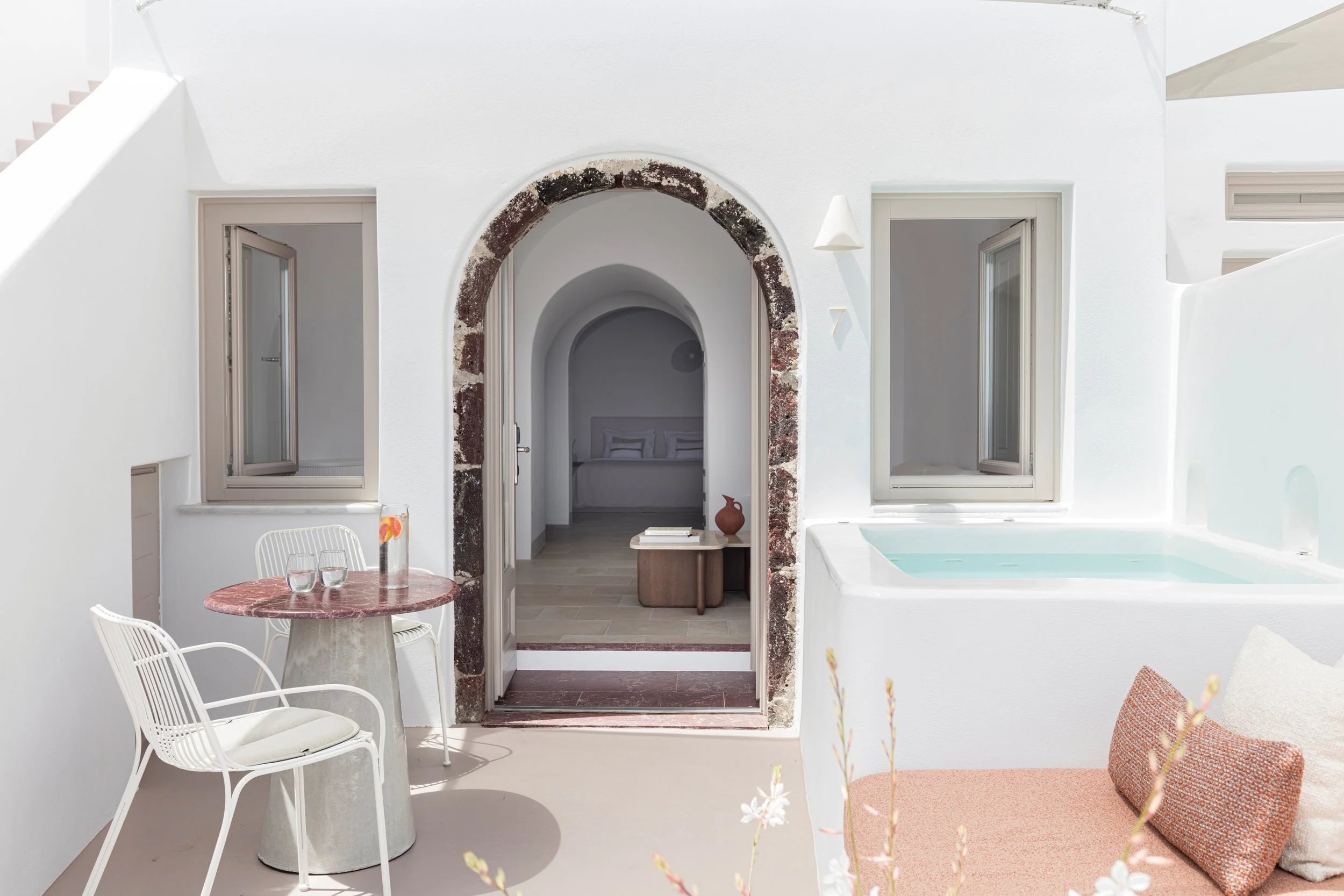 Outdoor patio area with white walls, a small round table with a pink marble top, two white wire chairs, a window with an open shutter, and a hot tub. An arched doorway with a rustic brick border leads to an indoor space where a bed with white linens 