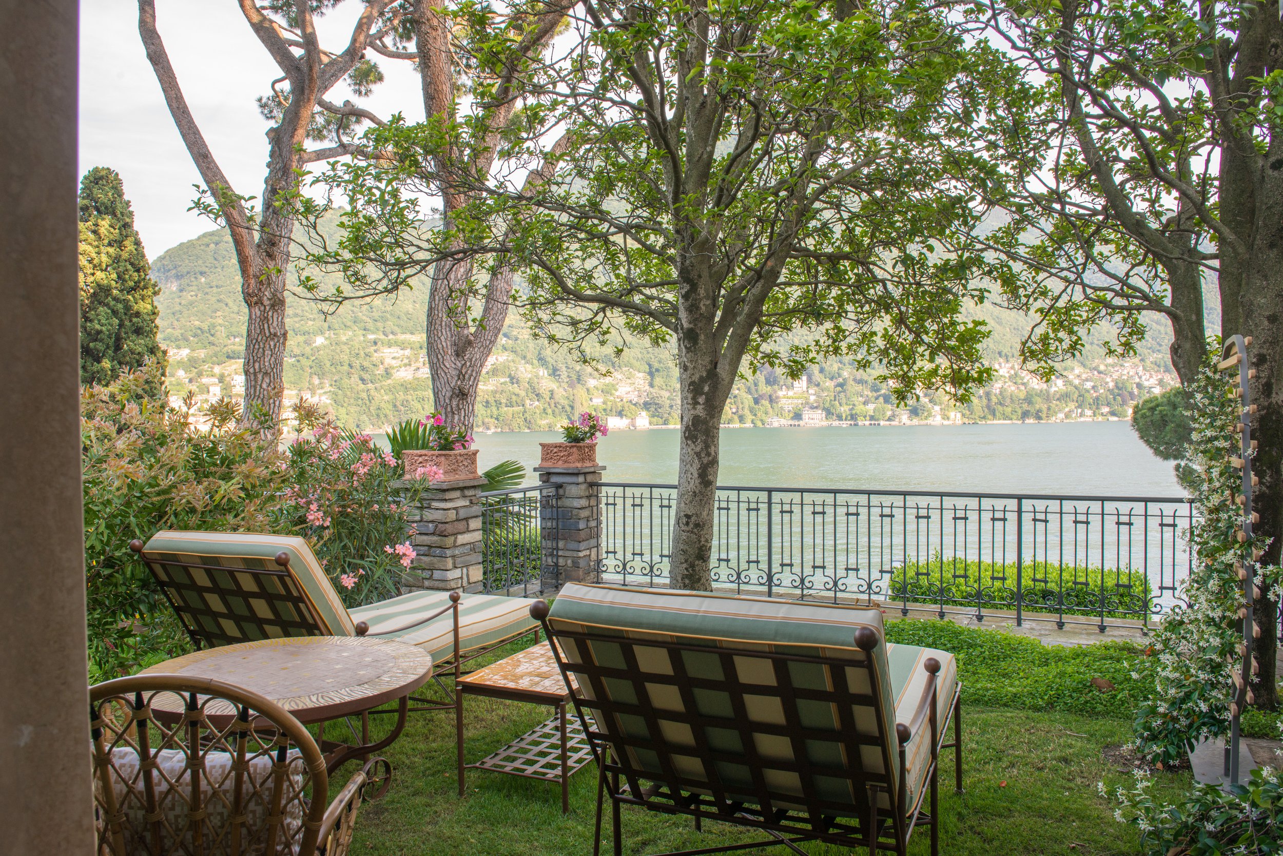 A lakeside patio with two lounge chairs, a small round table, and lush greenery, overlooking a lake and distant hills