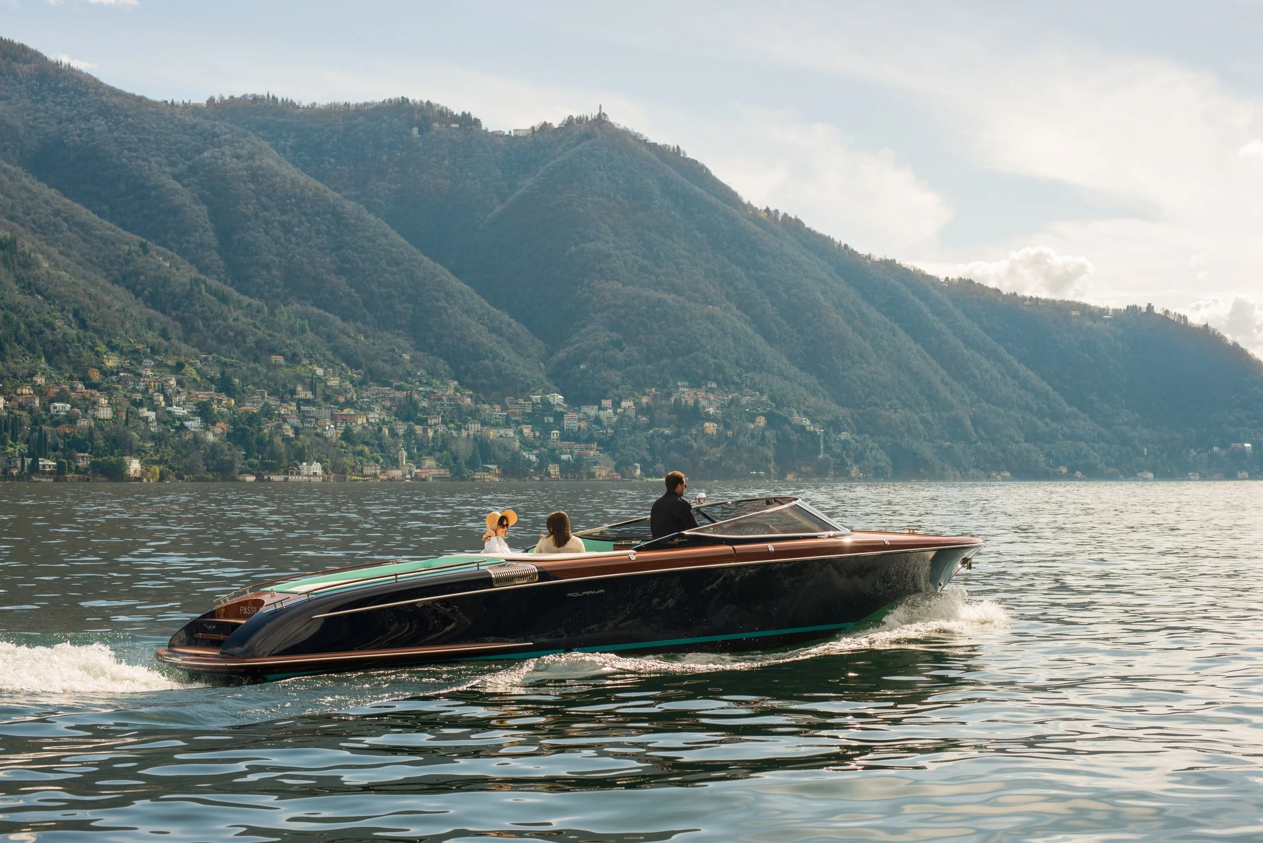 A luxury black speedboat with three people on board, cruising on a lake with a backdrop of green, mountainous terrain and scattered houses.