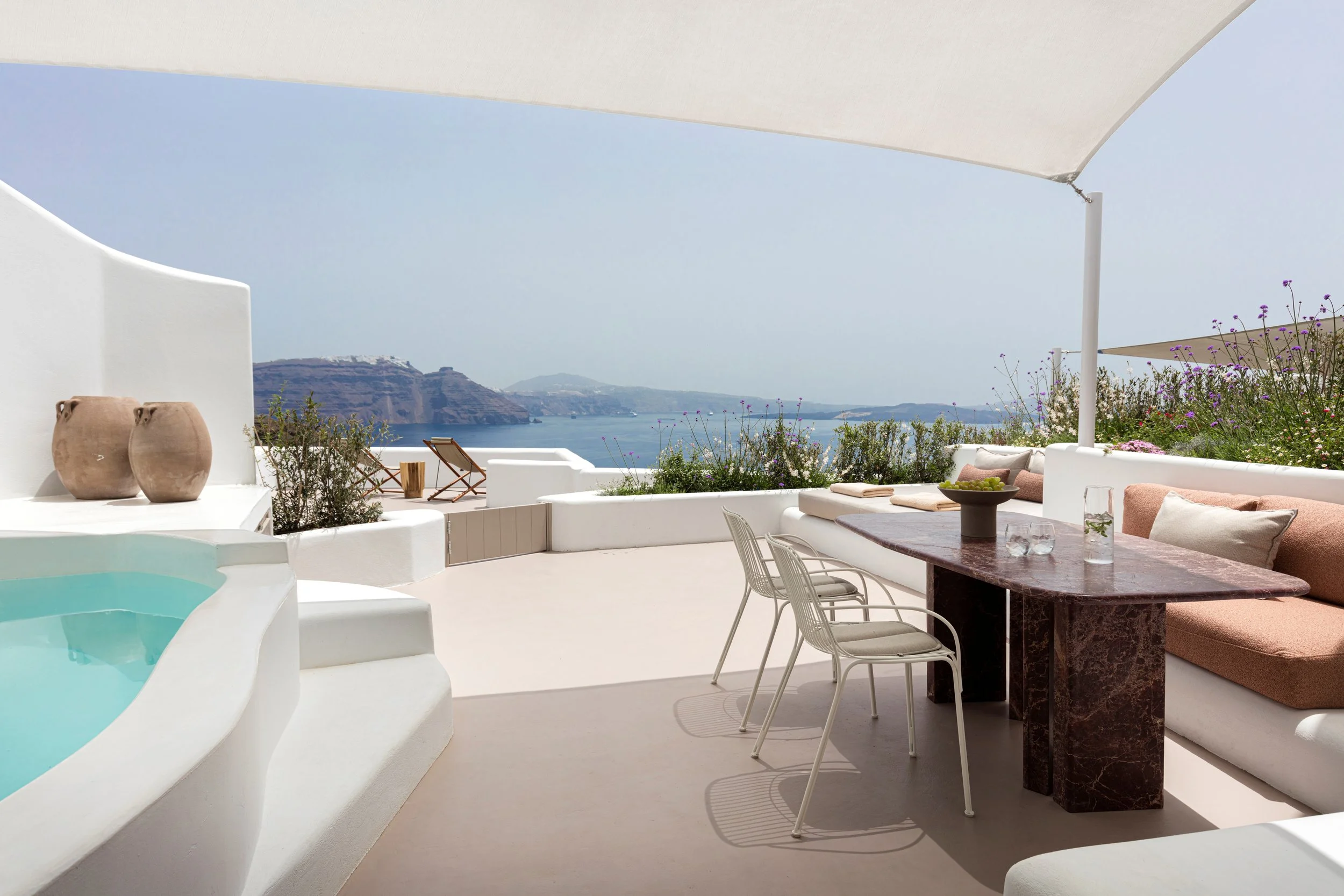 A luxurious outdoor terrace overlooking the ocean, with white walls, a hot tub, a dining table with chairs, and seating area with cushions and a garden bed with purple and pink flowers.
