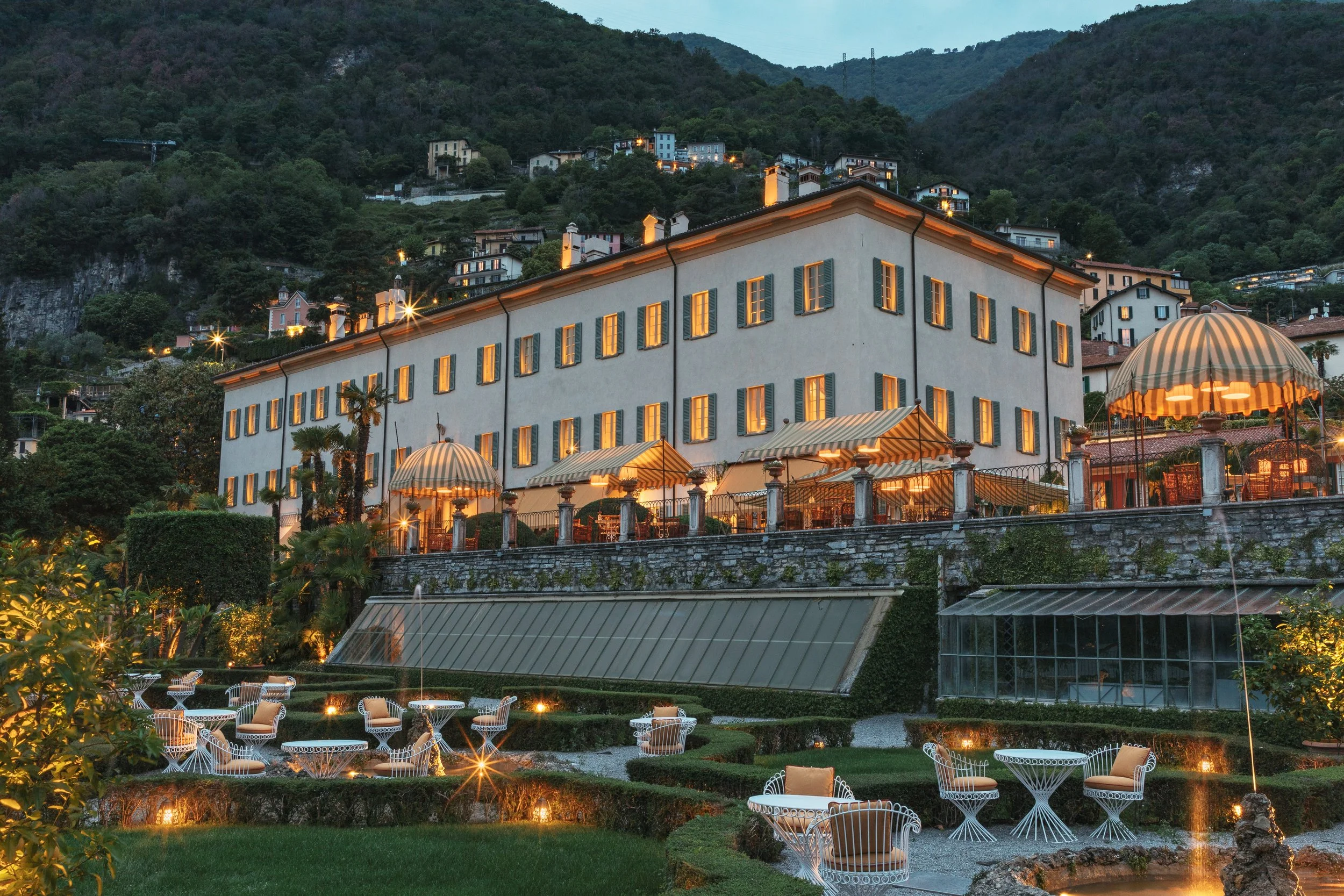 An elegant building with multiple windows and outdoor seating areas, situated on a hillside with lush greenery and other houses in the background during evening time.