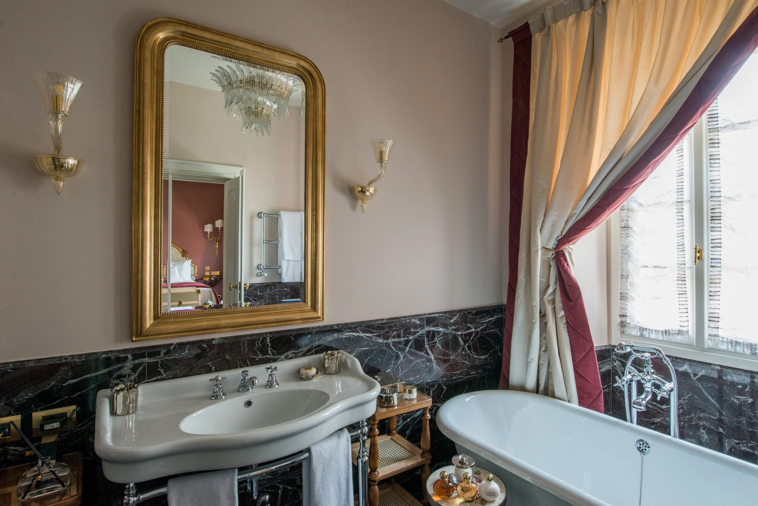 Luxury bathroom with a bathtub, sink, mirror, elegant wall sconces, and a large window with draped curtains.