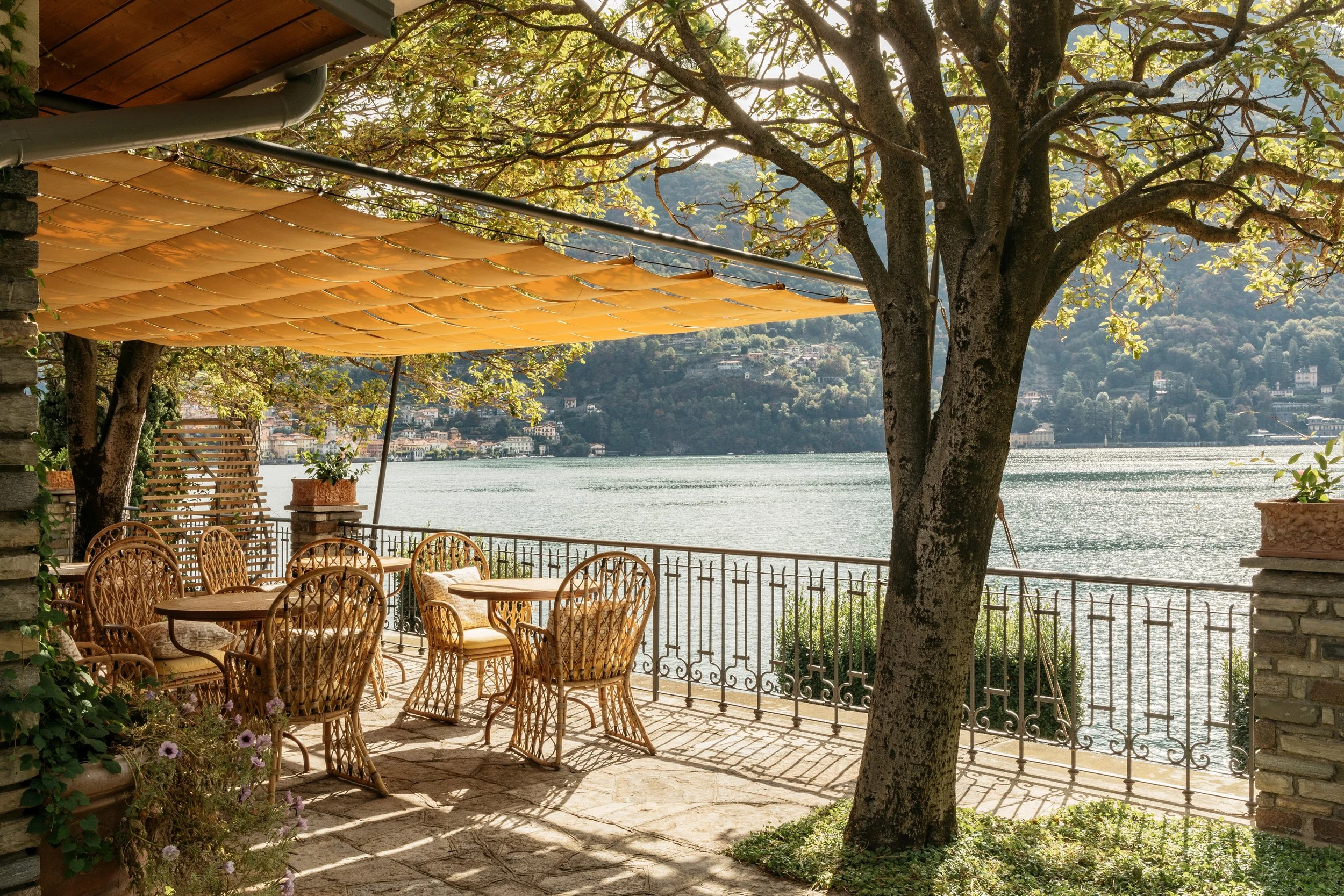 A lakeside outdoor patio with wicker chairs and tables, shaded by a tree and a yellow canopy, overlooking a body of water with hills and houses in the distance.