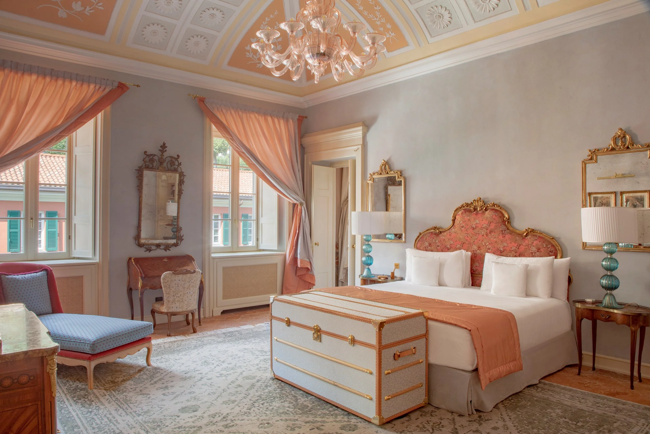 Elegant hotel bedroom with pastel-colored walls, a large bed with a pink and beige headboard, and vintage furniture including bedside tables with lamps, a bench at the foot of the bed, and a seating area with chairs and a small desk. Light streams through the window with pink curtains.
