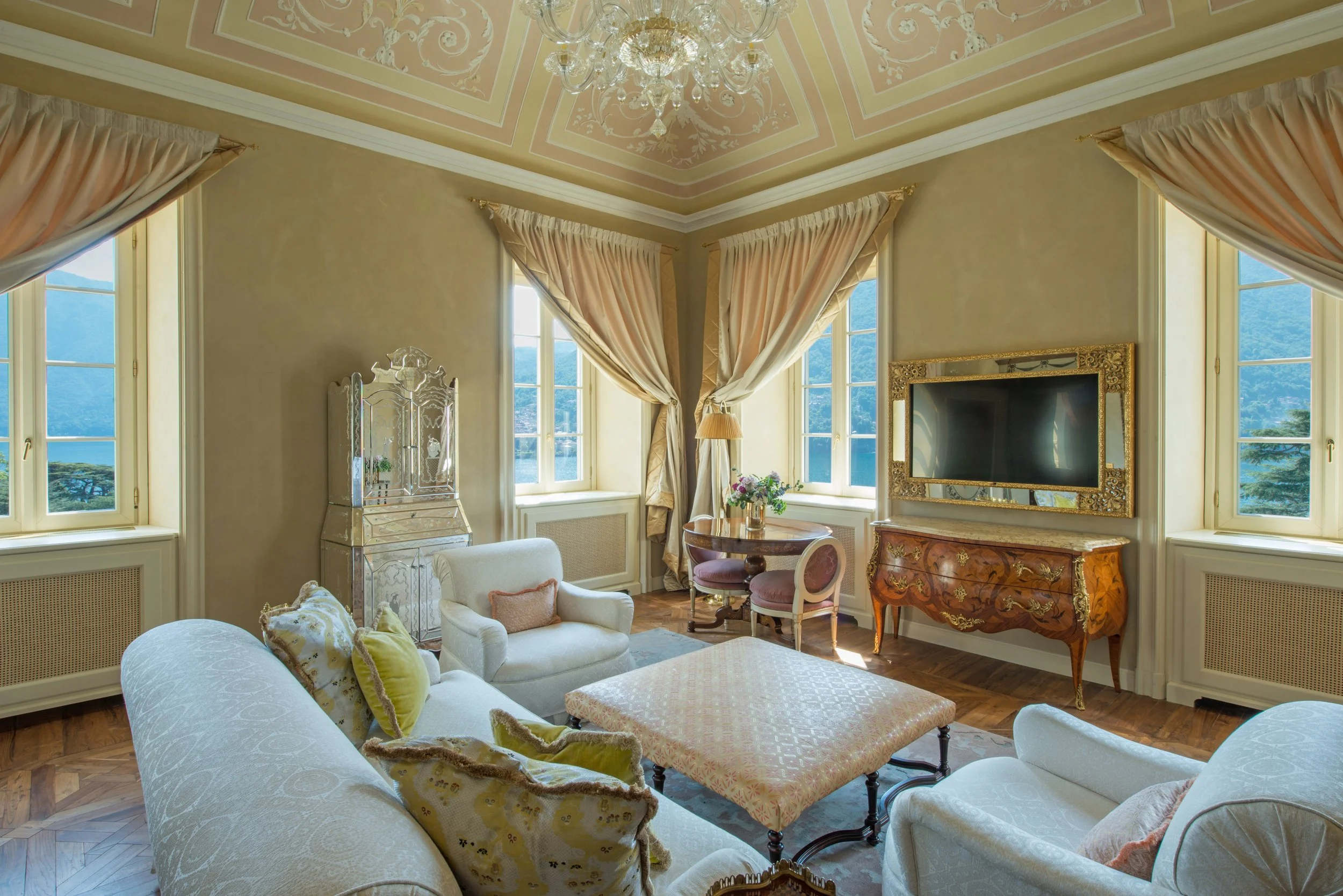 Elegant living room with cream walls, multiple large windows with beige curtains, a chandelier, a flat-screen TV in an ornate gold frame, a wooden chest, upholstered armchairs and a sofa with decorative pillows, a diamond-patterned coffee table, and a small round dining table with purple chairs, overlooking a landscape of mountains and trees outside the windows.