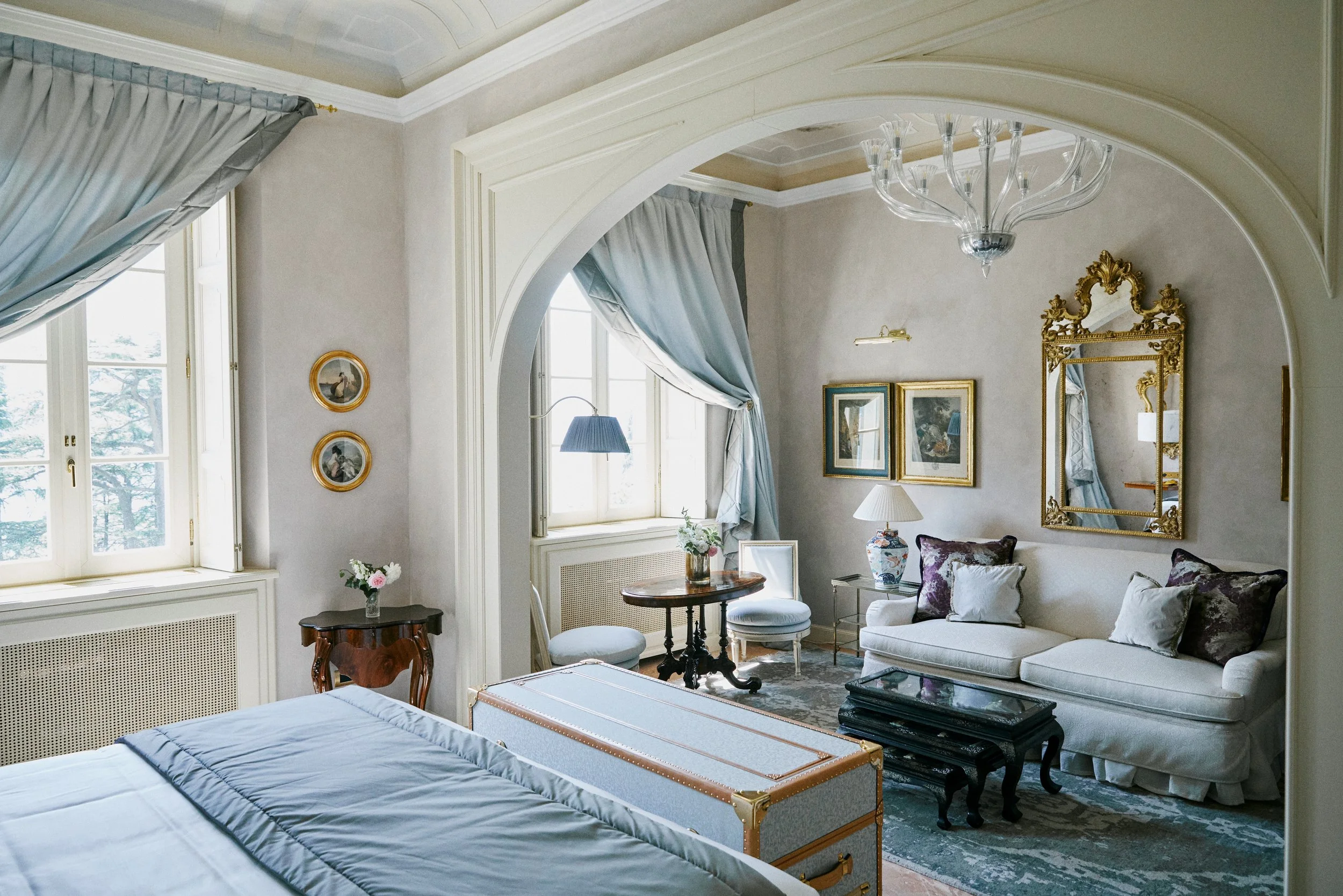 Elegant hotel room with a bed, a sitting area with a white sofa, pillows, and a small round table near windows with draped curtains. Decor includes a large ornate mirror, framed pictures, lamps, and floral arrangements.