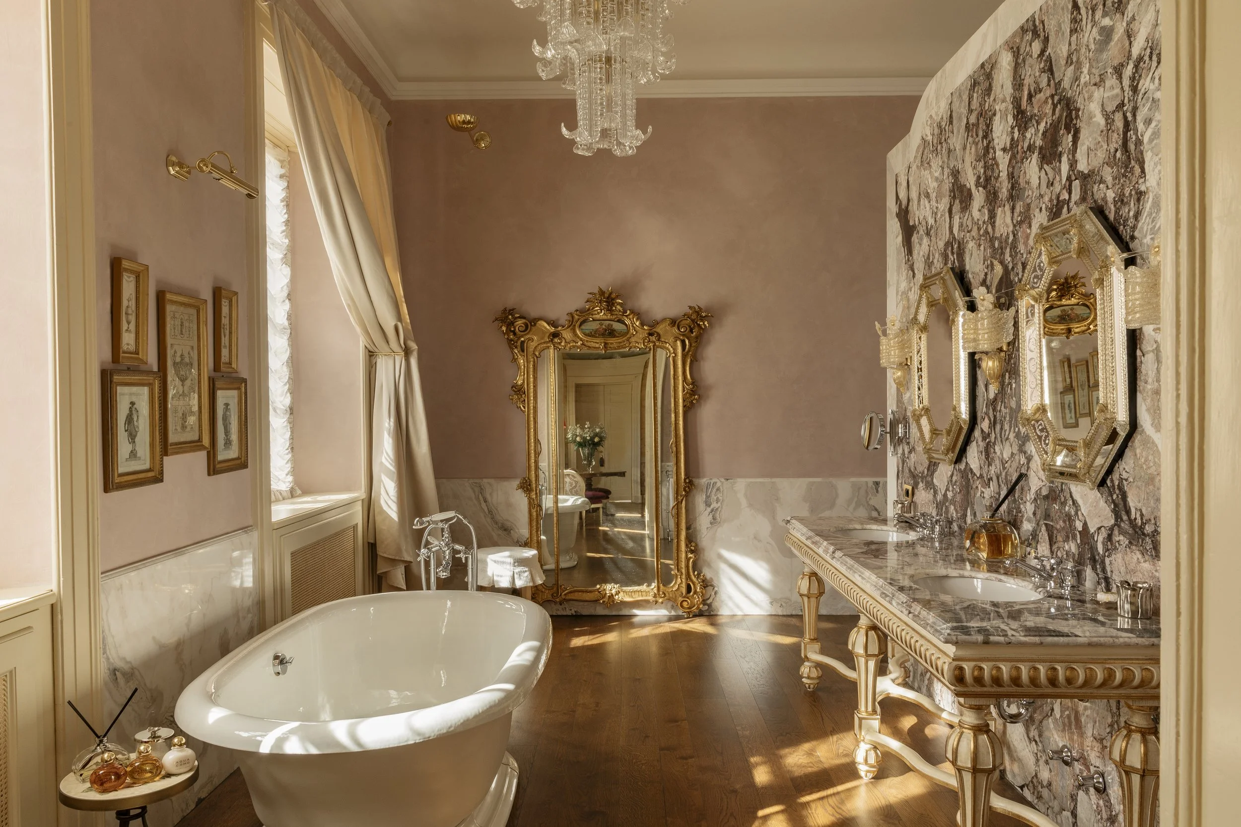 Luxurious bathroom with a clawfoot bathtub, marble countertop double sinks, ornate gold-framed mirror, decorative wall mirrors, framed artwork, large window with drapes, chandelier, and hardwood floor.