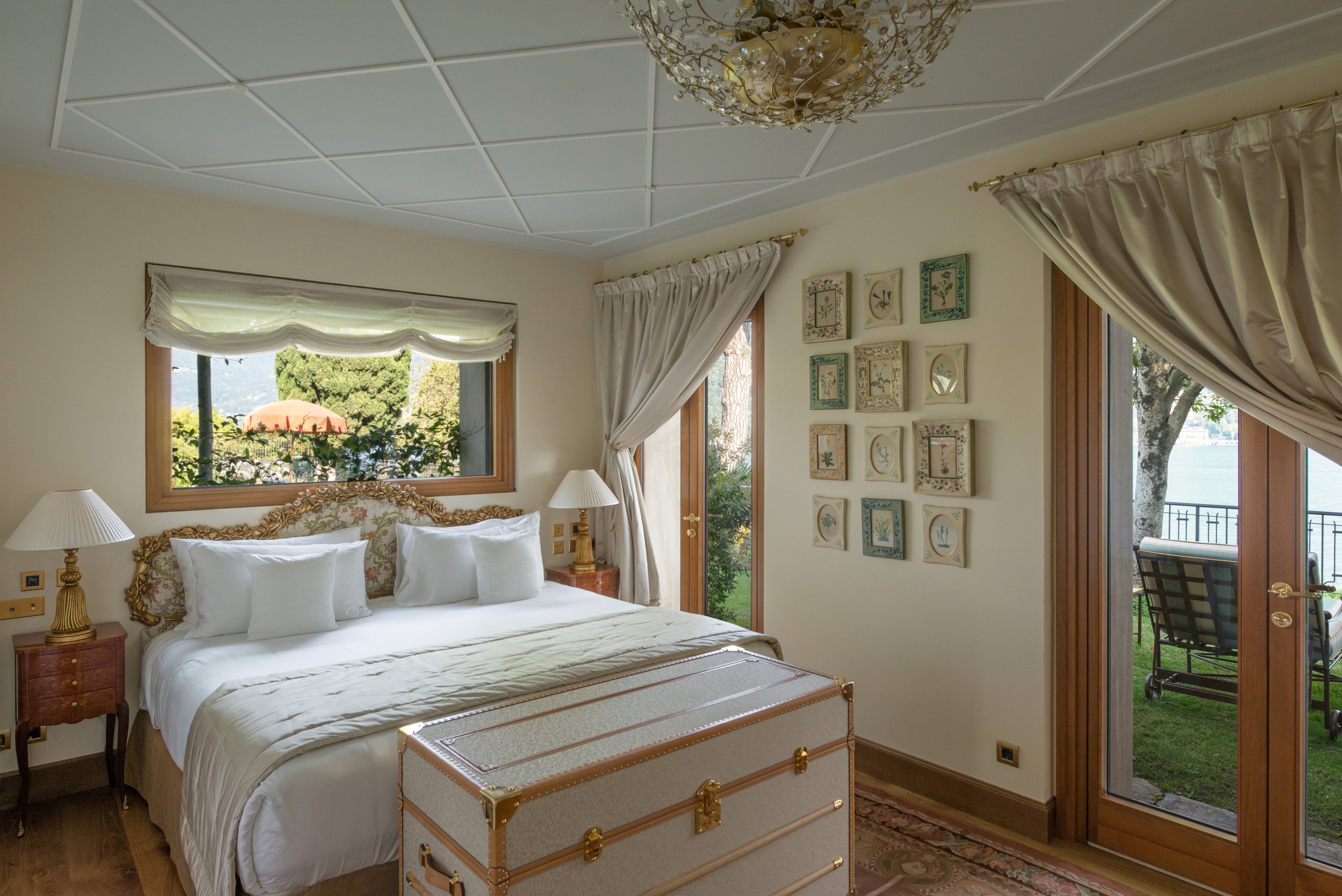 A cozy bedroom with a large bed decorated with ornate gold and fabric headboard, white bedding, and multiple pillows. There are bedside tables with matching lamps on each side. The room has wooden floors, cream-colored walls, and large windows and a glass door with curtains, revealing an outdoor patio or garden area with chairs and a view of water and trees.