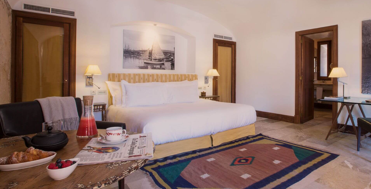 A hotel room with a king-size bed, reading lamps, artwork of sailboats, a wooden table with breakfast items, and a colorful rug.