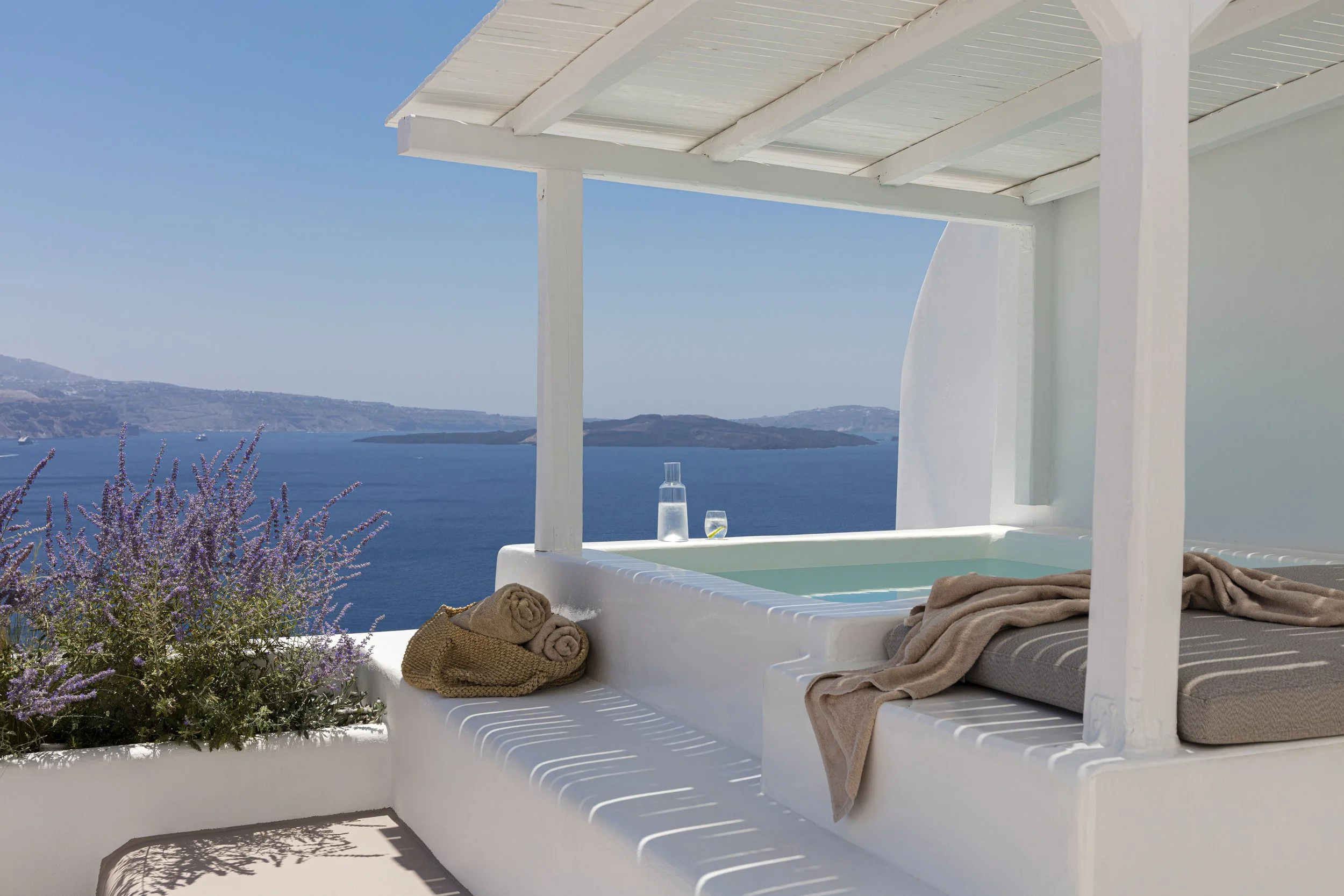 A rooftop hot tub with a view of the ocean and islands, decorated with towels, a water bottle, a glass of water, and purple flowers in a white planter.