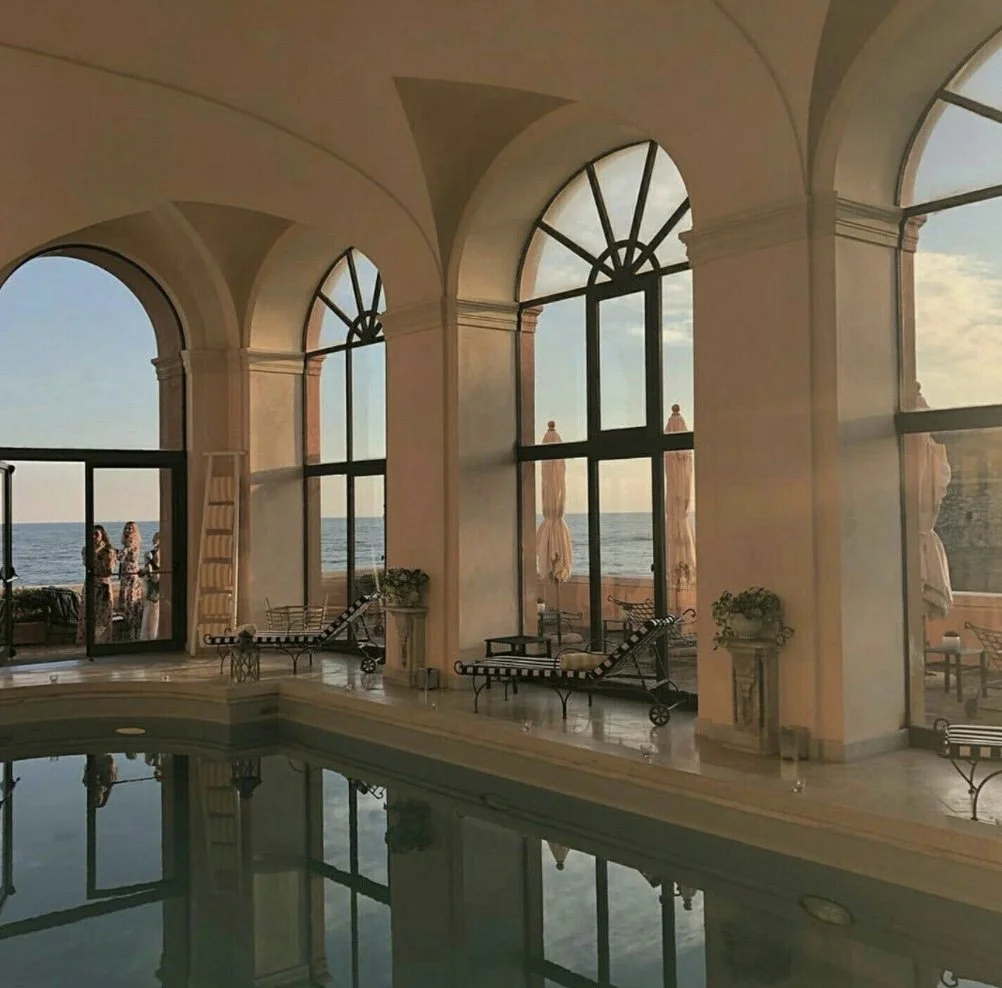 Indoor pool area with large arched windows overlooking the ocean, two lounge chairs, a ladder, and several people near the windows enjoying the view.