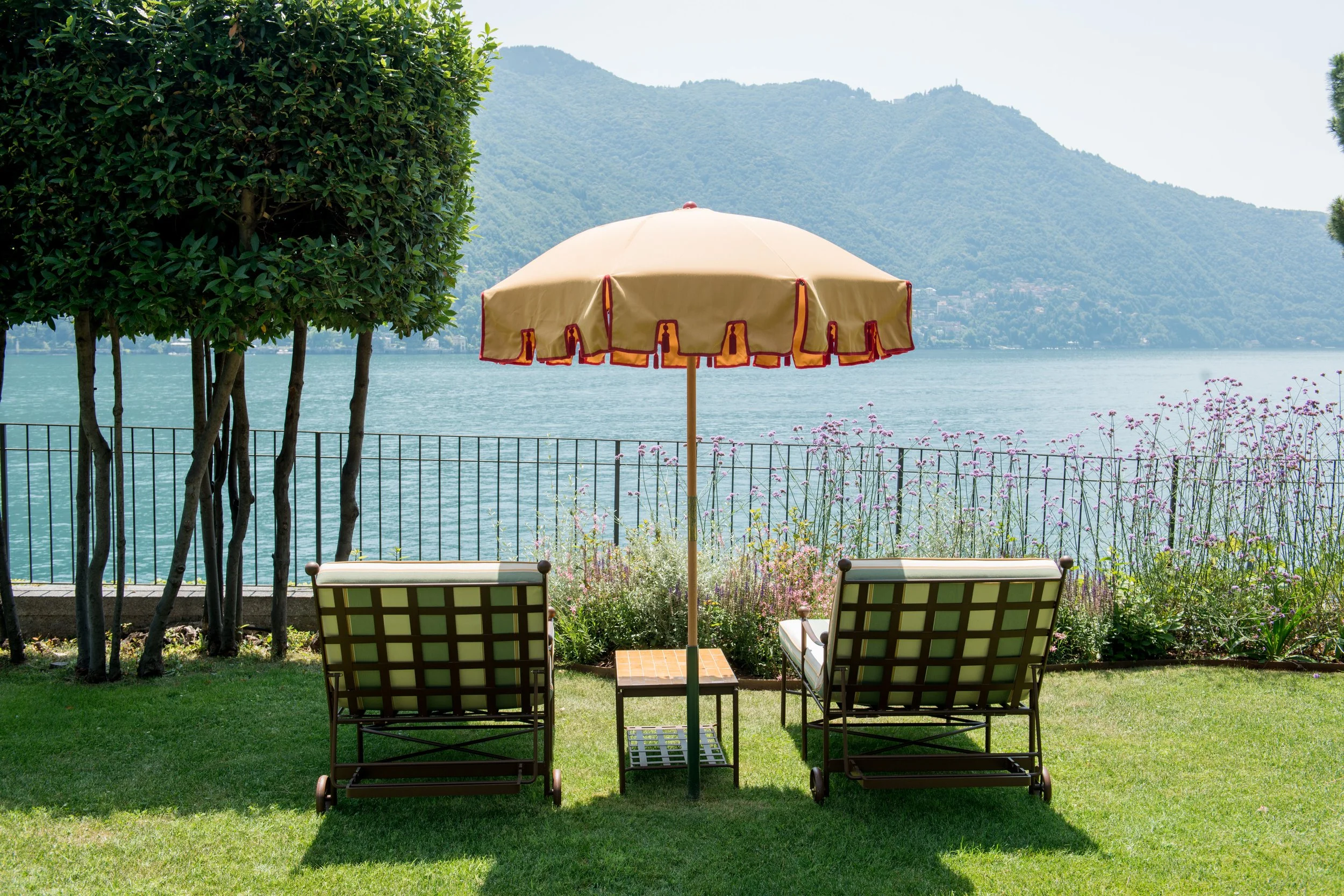 Two lounge chairs with a small table and an umbrella on a grassy area overlooking a lake with mountains in the background.