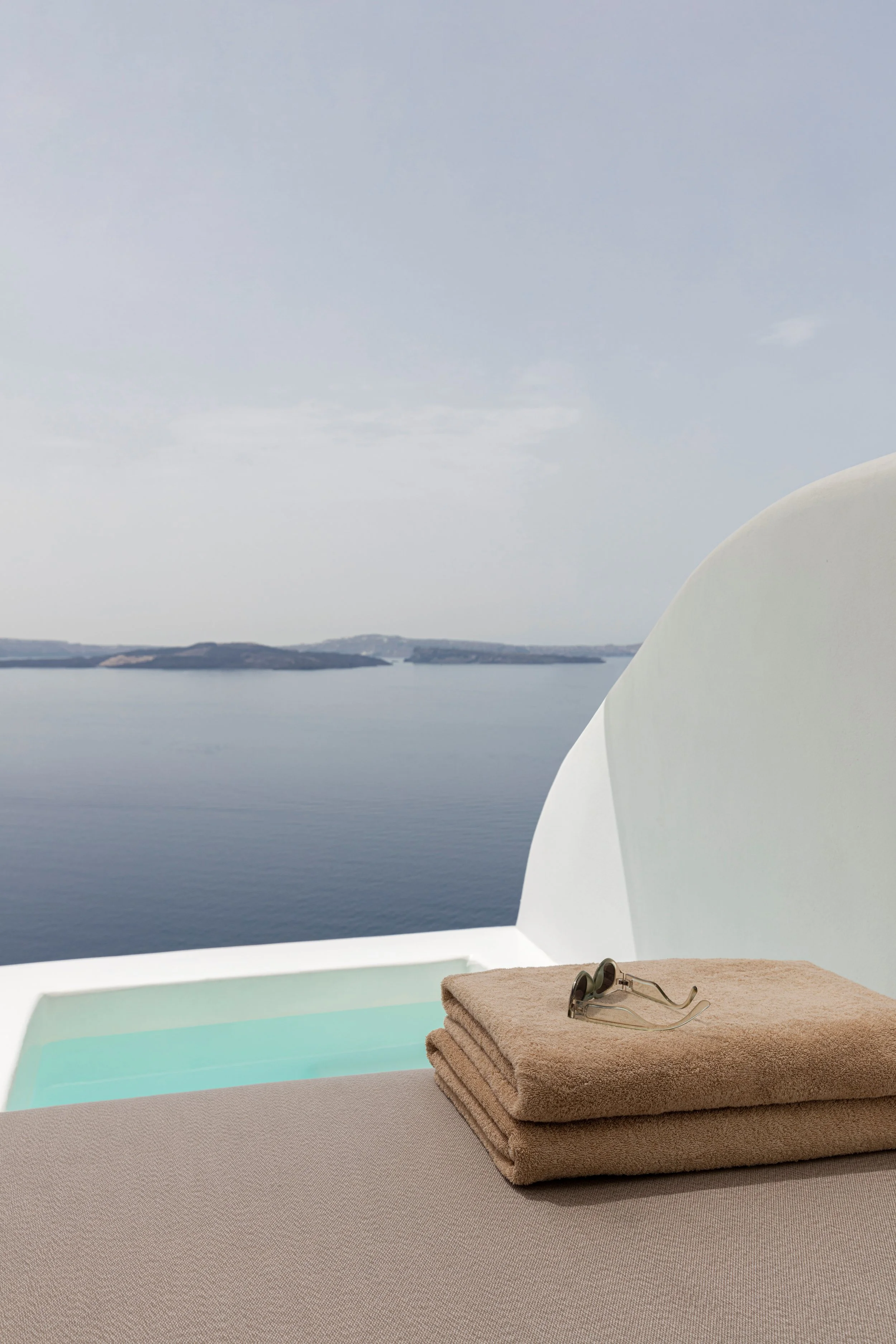 View of a white bed or lounger with folded towels and sunglasses, overlooking a body of water and distant islands or landforms.