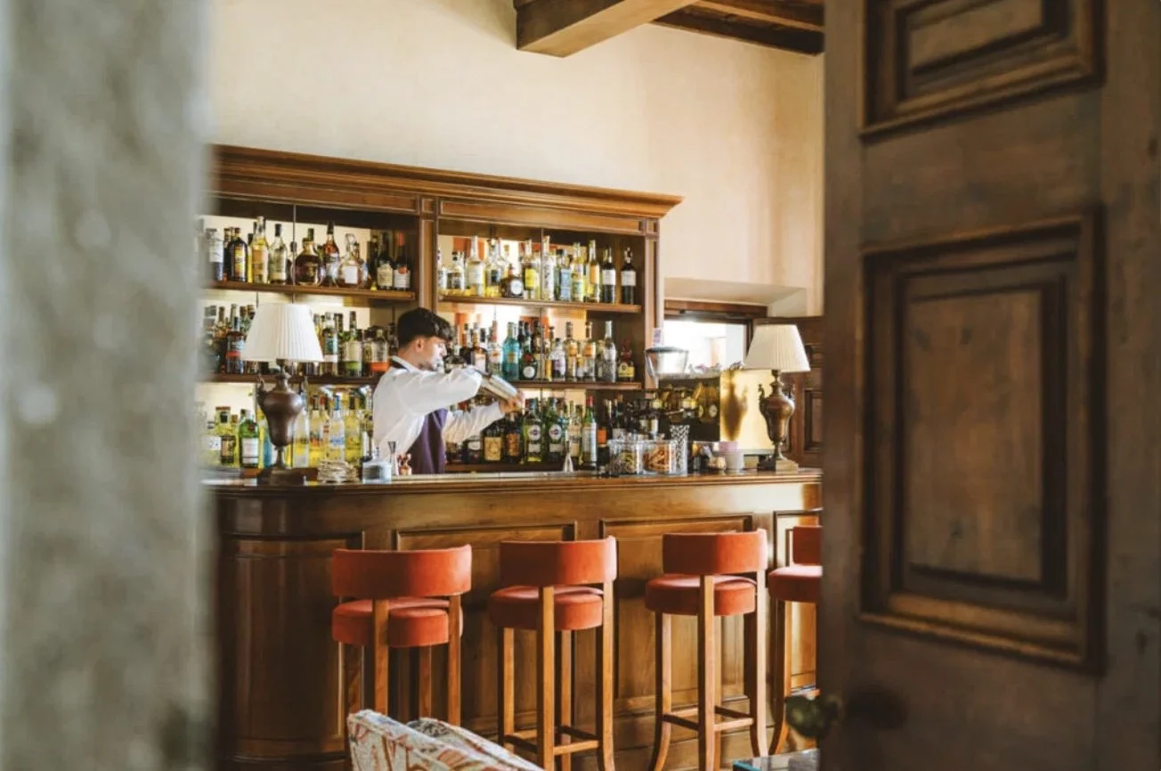 A bartender mixing drinks behind a wooden bar with shelves of liquor bottles, two lamps, and four red bar stools in front of the bar.