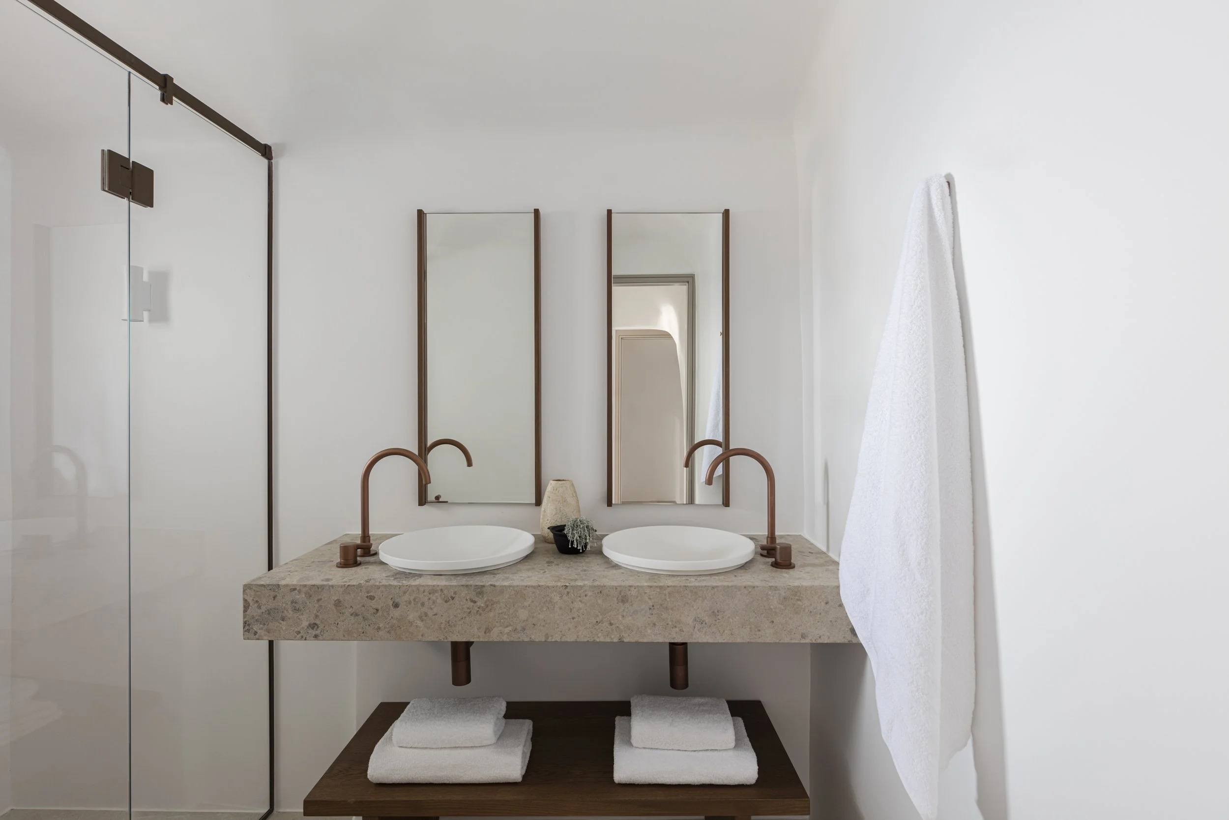 Modern bathroom with double sink vanity, wall-mounted mirrors, copper fixtures, white towels, and a glass shower door.