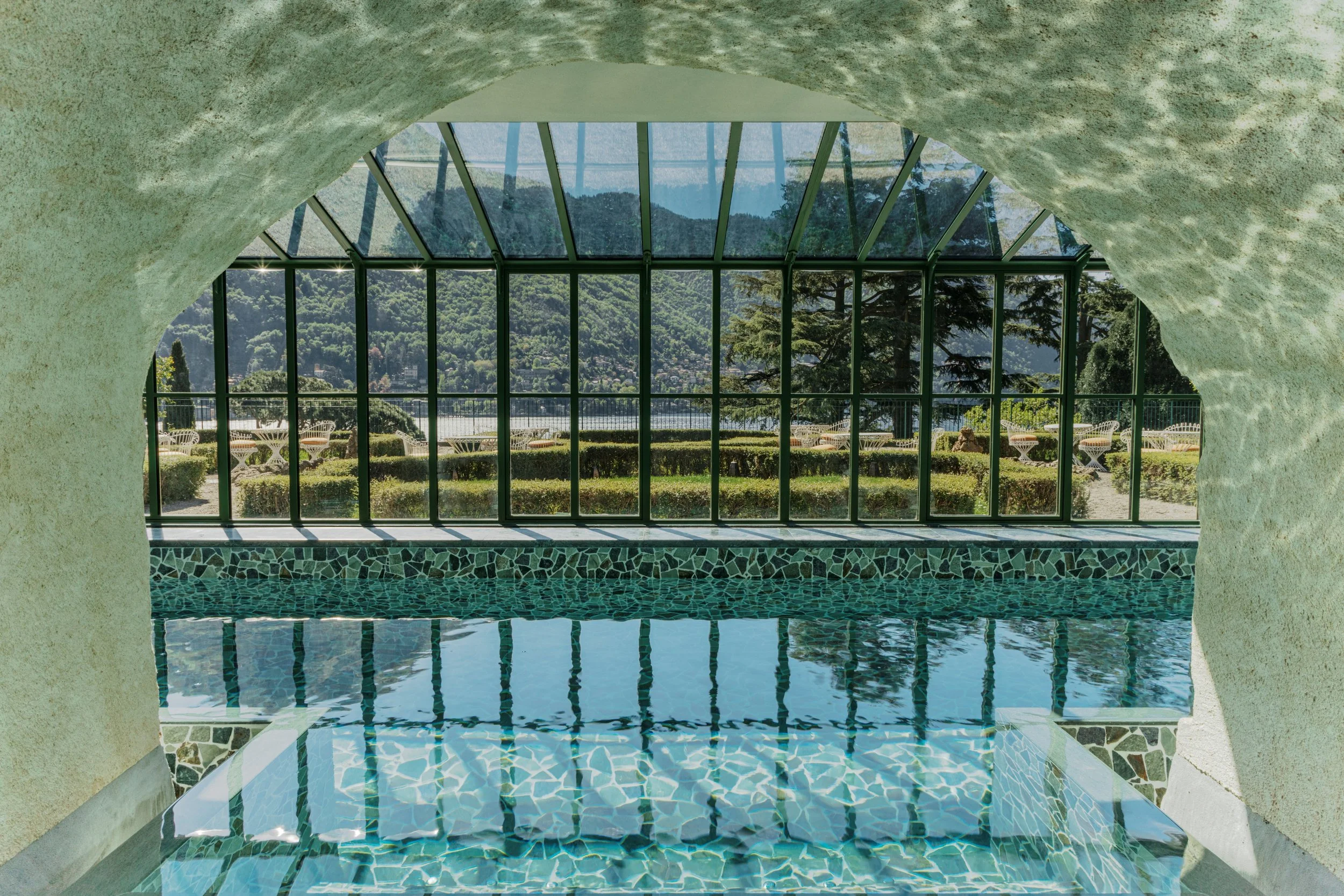 Interior view of a swimming pool with mosaic tiles, looking through a large glass window externally showing outdoor landscape with trees, hills, and a sunny day.