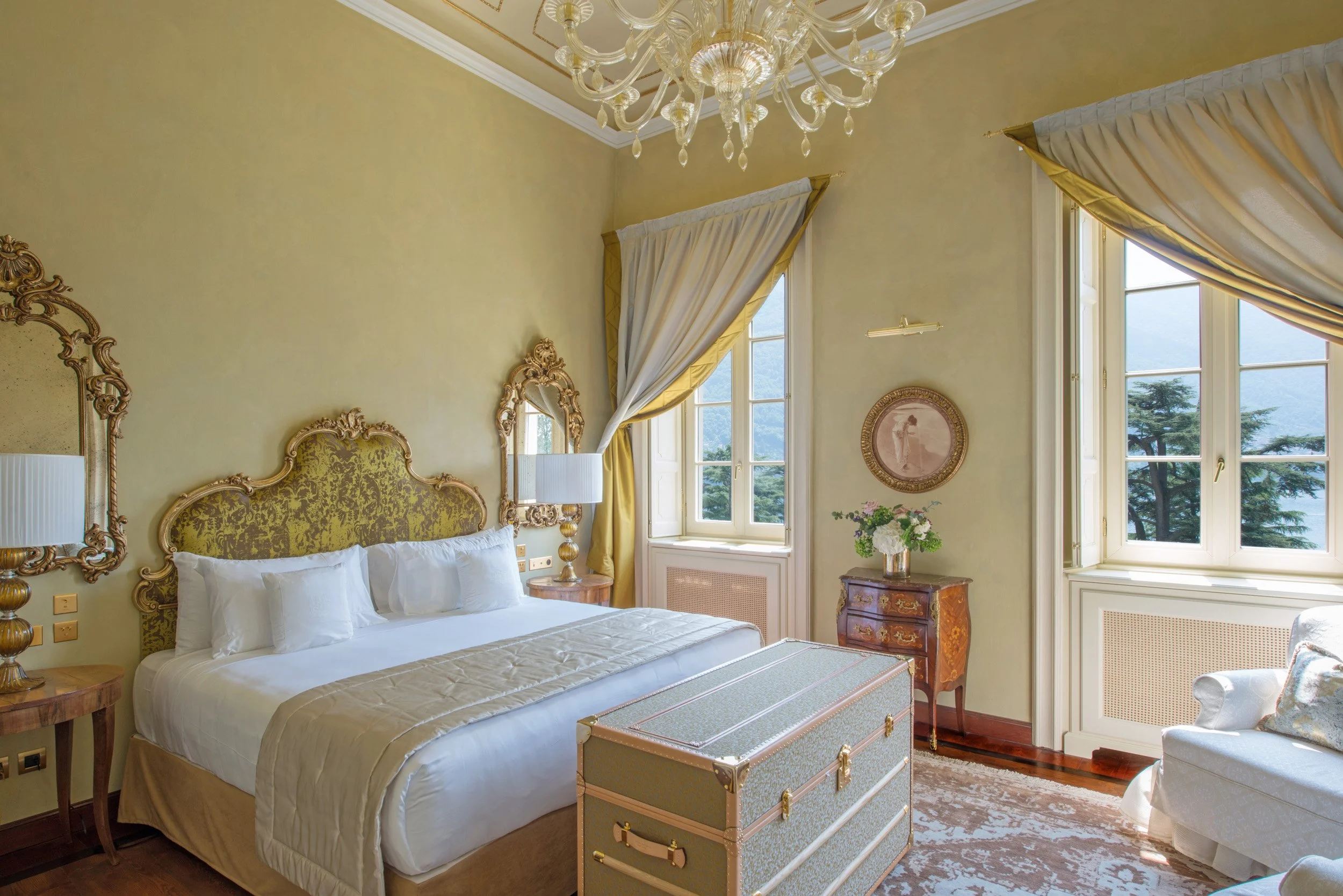 A luxurious bedroom with a large bed with a gold ornate headboard, two side tables with lamps, and two windows with heavy draped curtains. The room has a chandelier and vintage furniture, including a chest at the foot of the bed and a small table with a vase of flowers.