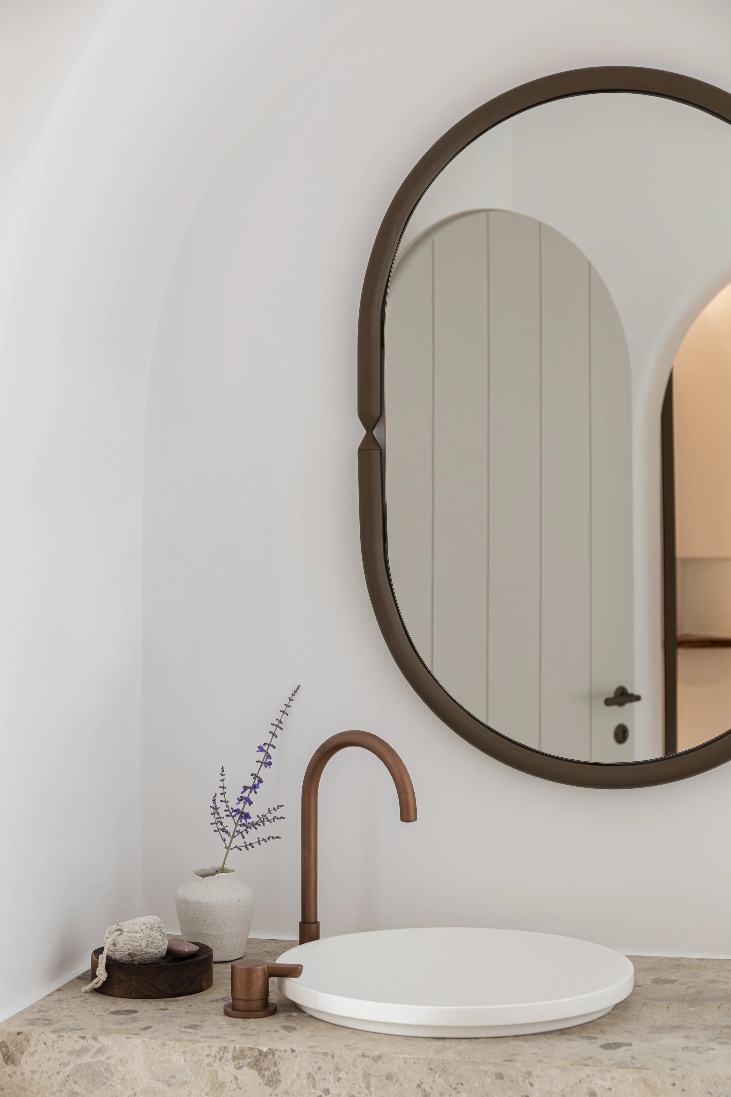 A modern bathroom sink with a round mirror, a copper faucet, a small potted lavender plant, and decorative stones on a marble countertop.