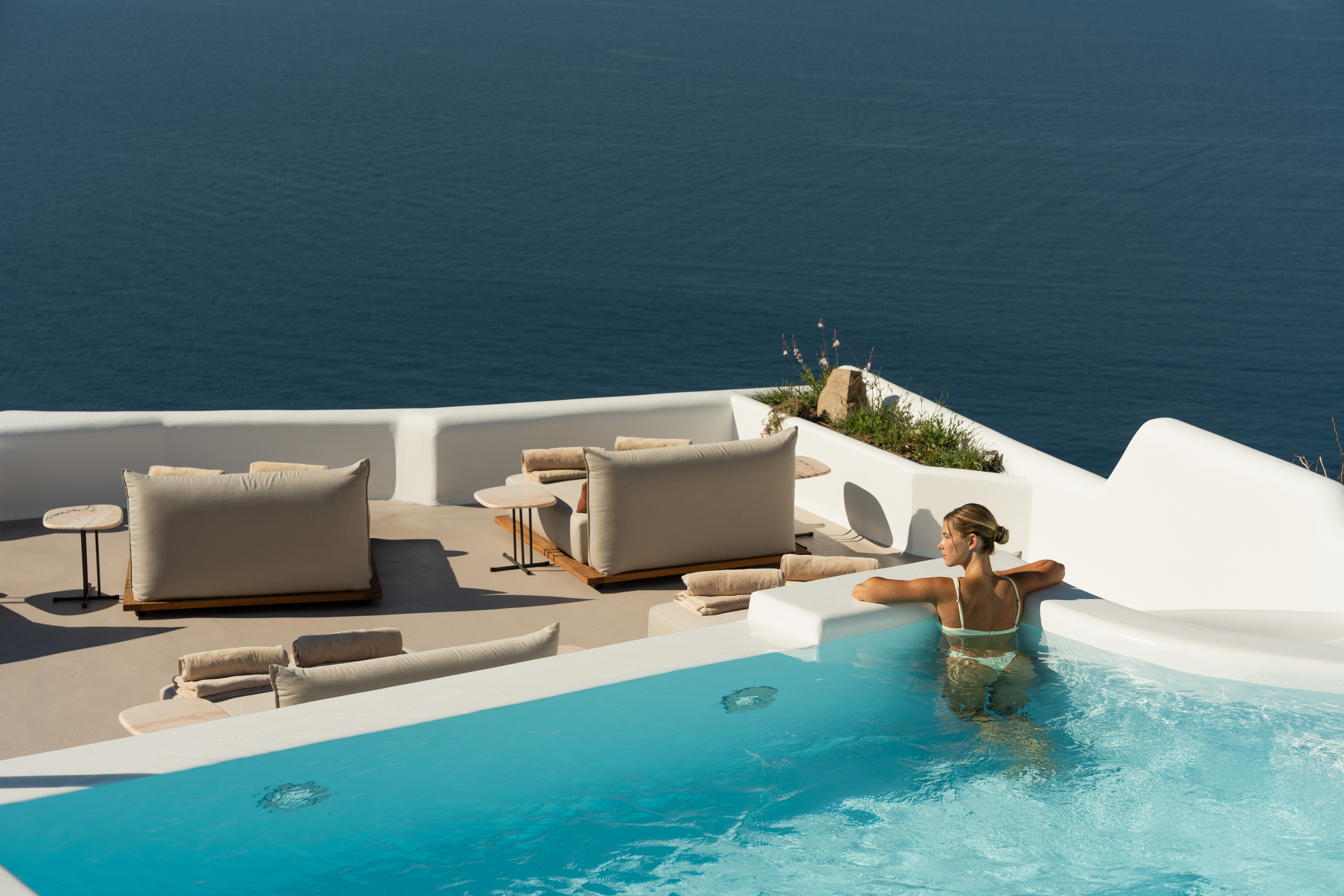 A woman relaxing in a hot tub near the ocean with outdoor seating and white architecture.