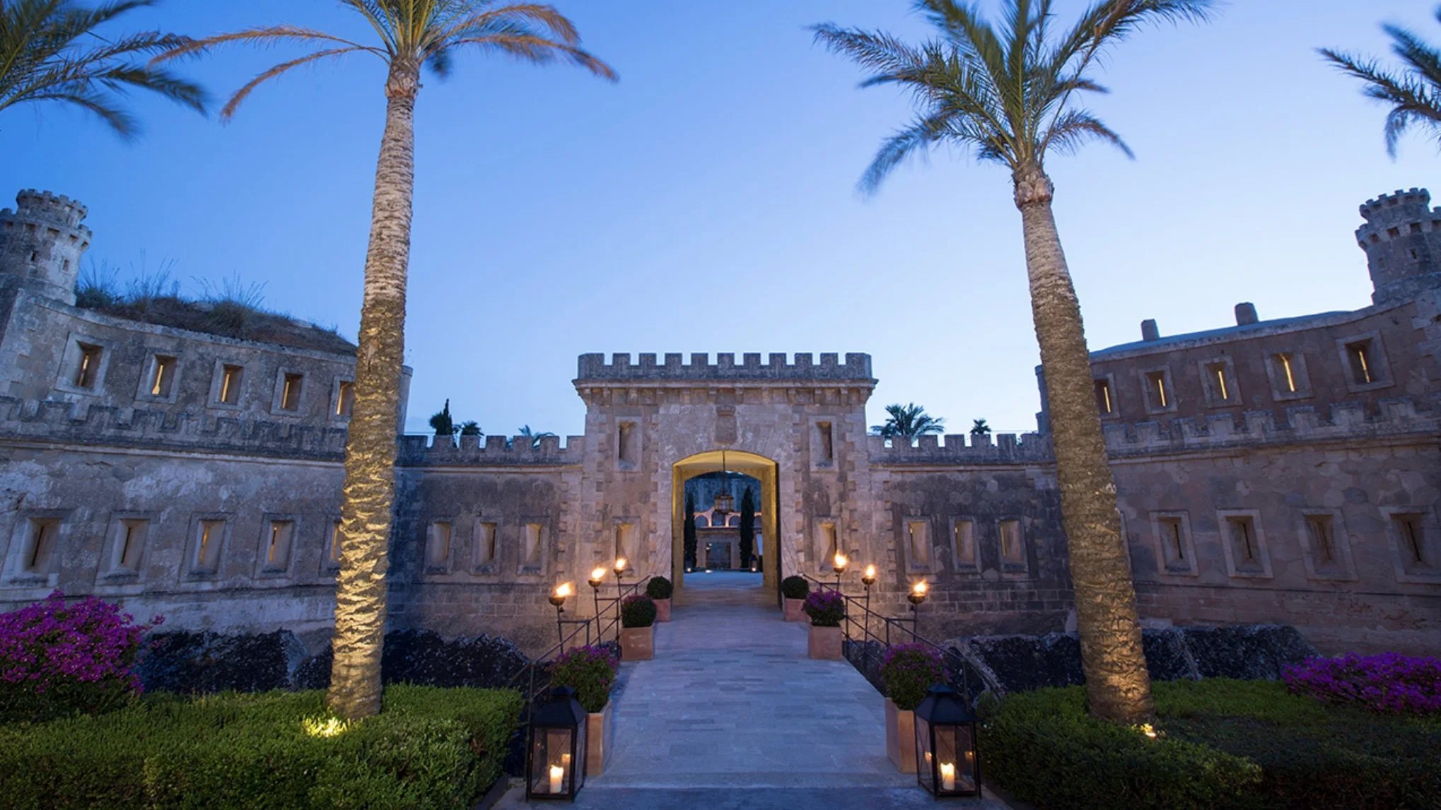 A castle-style stone entrance with towers on each side, palm trees on either side of the pathway, and lit lanterns lining the walkway during dusk.