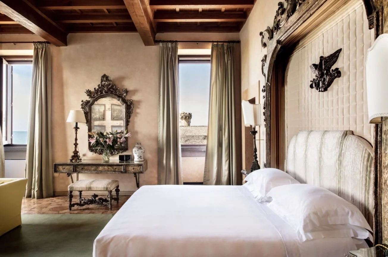A luxurious bedroom with a large bed, ornate wooden headboard, and vintage decor, including lamps, a mirror, and a vase of fresh flowers, with windows revealing the outdoors.