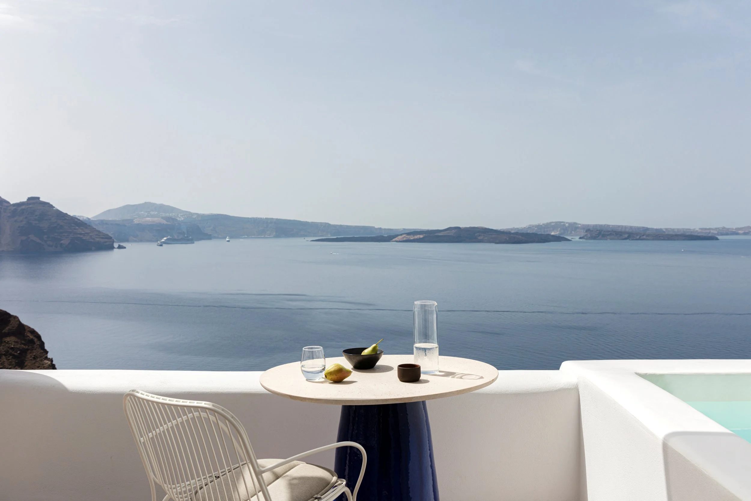 A white circular table with a black bowl of pears, a glass of water, a clear water pitcher, and a small brown cup on a blue base, along with a white chair, overlooking a calm sea with islands and boats in the distance on a bright day.