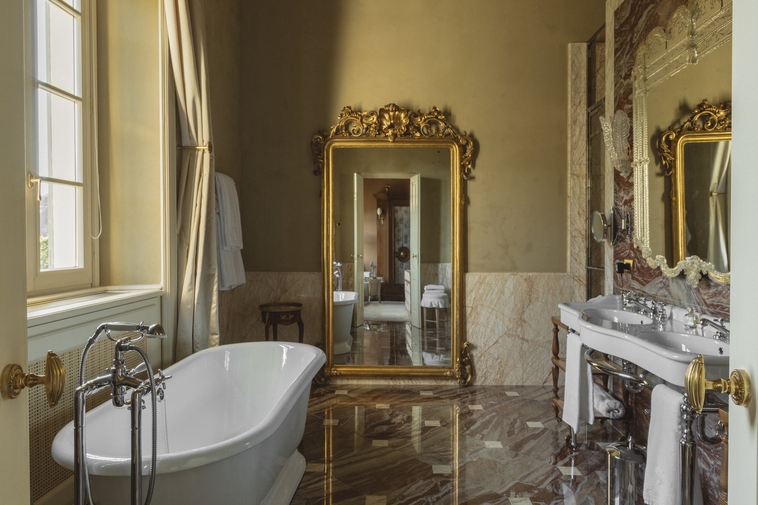 Luxurious bathroom with a clawfoot bathtub, a large ornate gold-framed mirror, double sinks with decorative mirror, marble floors, and elegant window drapes.