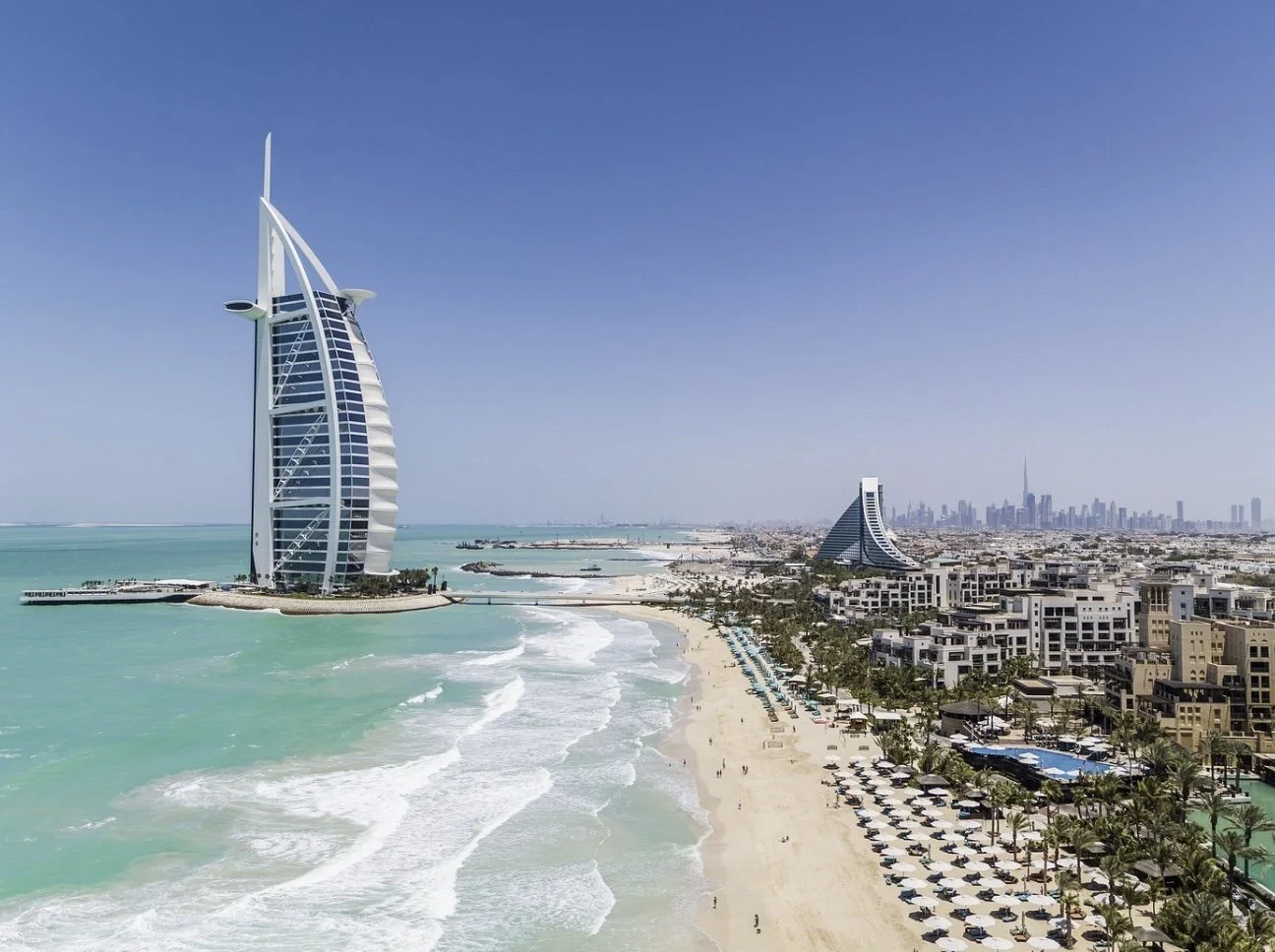Aerial view of Dubai coastline featuring the Burj Al Arab hotel, sandy beach with umbrellas, high-rise buildings, and the Dubai skyline in the background.