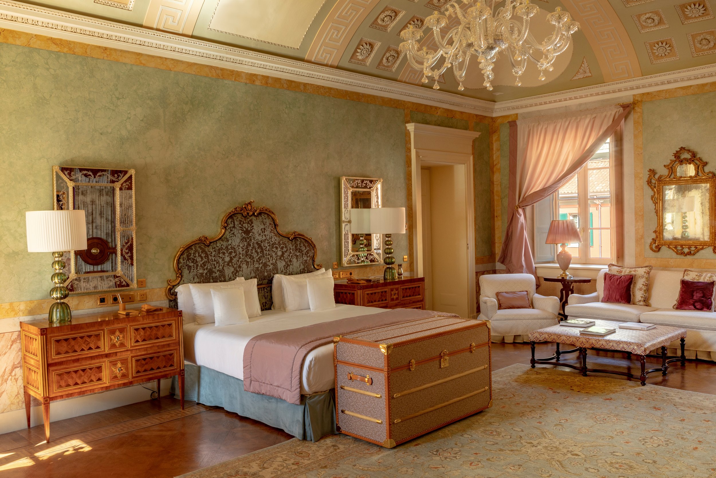 Luxurious hotel room with a large bed, two matching bedside tables with lamps, a sitting area with armchairs and a sofa, and a chandelier hanging from a decorated ceiling.