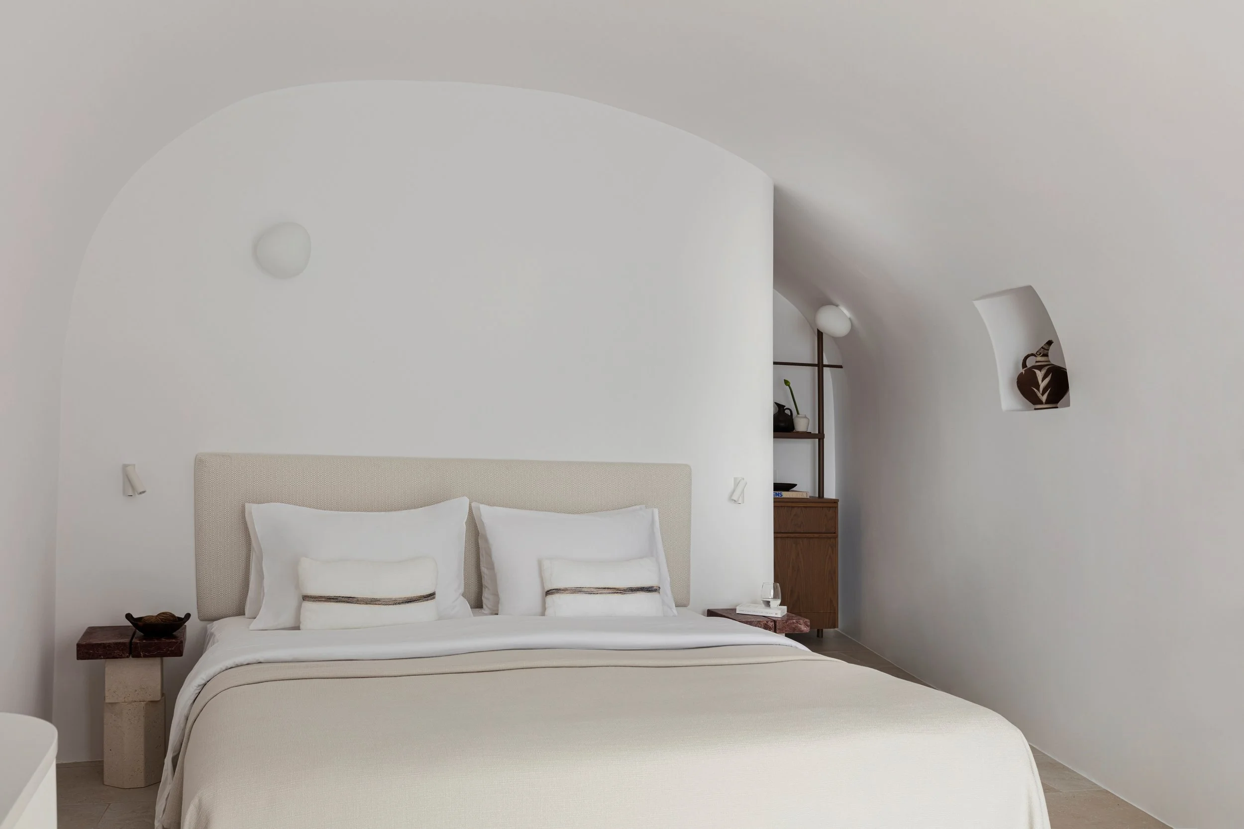 A minimalist bedroom with a white bed, beige headboard, and white bedding. There are two pillows and a small decorative pillow with a simple stripe. Side tables on each side of the bed, with a glass of water on the right table. The room has rounded w