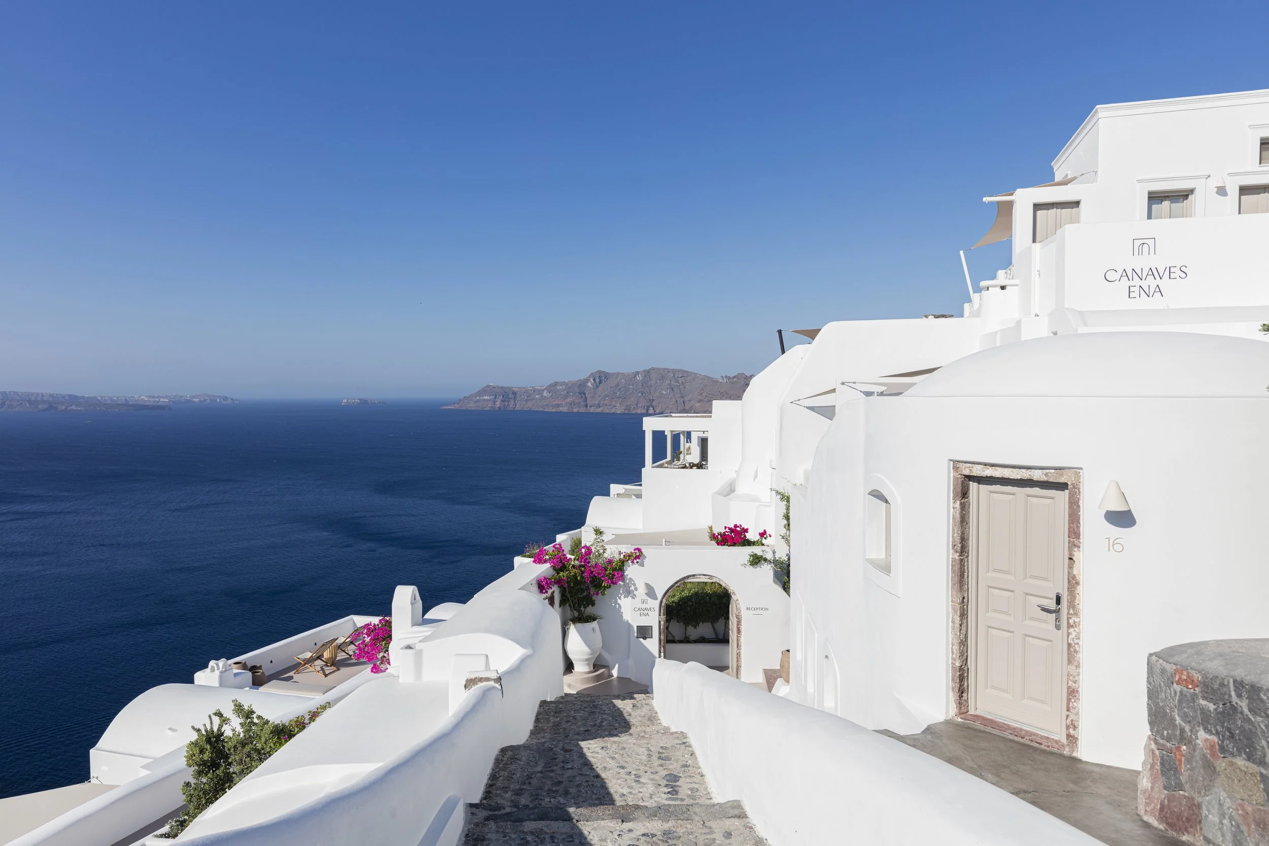 White buildings with pink flowers on a hillside overlooking the deep blue ocean in Santorini, Greece.
