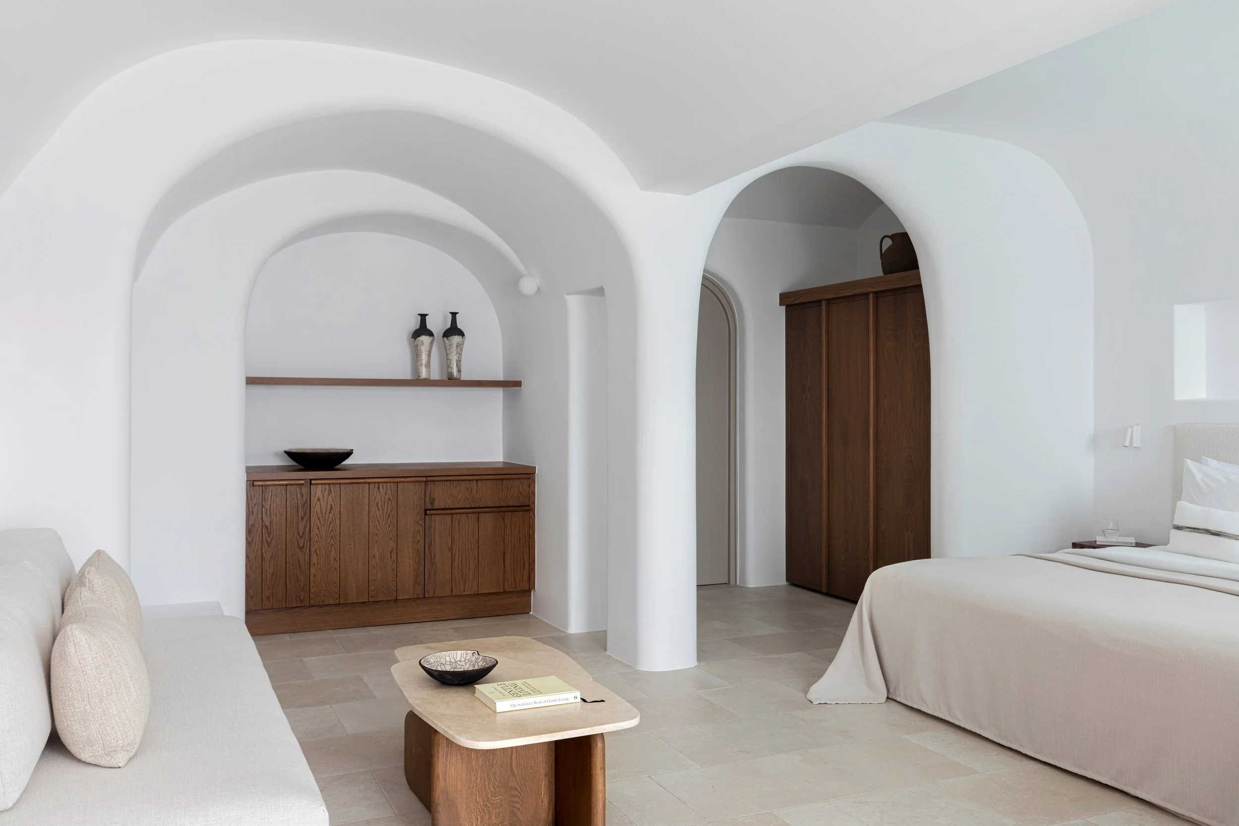Minimalist hotel room with white walls, arched doorway, wooden furniture, a bed with white bedding, beige sofa, and a small wooden coffee table.