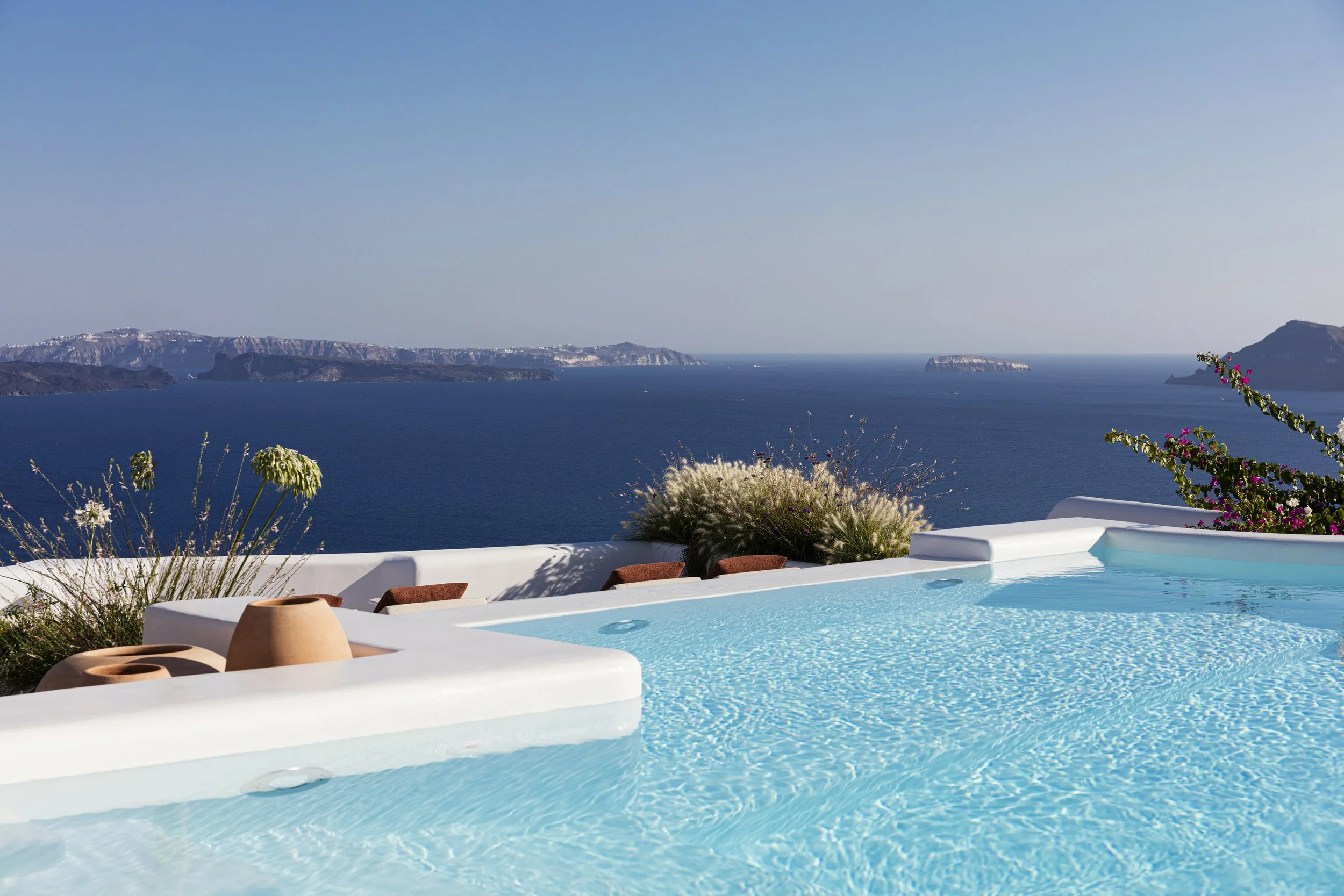 Infinity pool with a view of the ocean and mountains, white walls, potted plants, and flowering shrubs.
