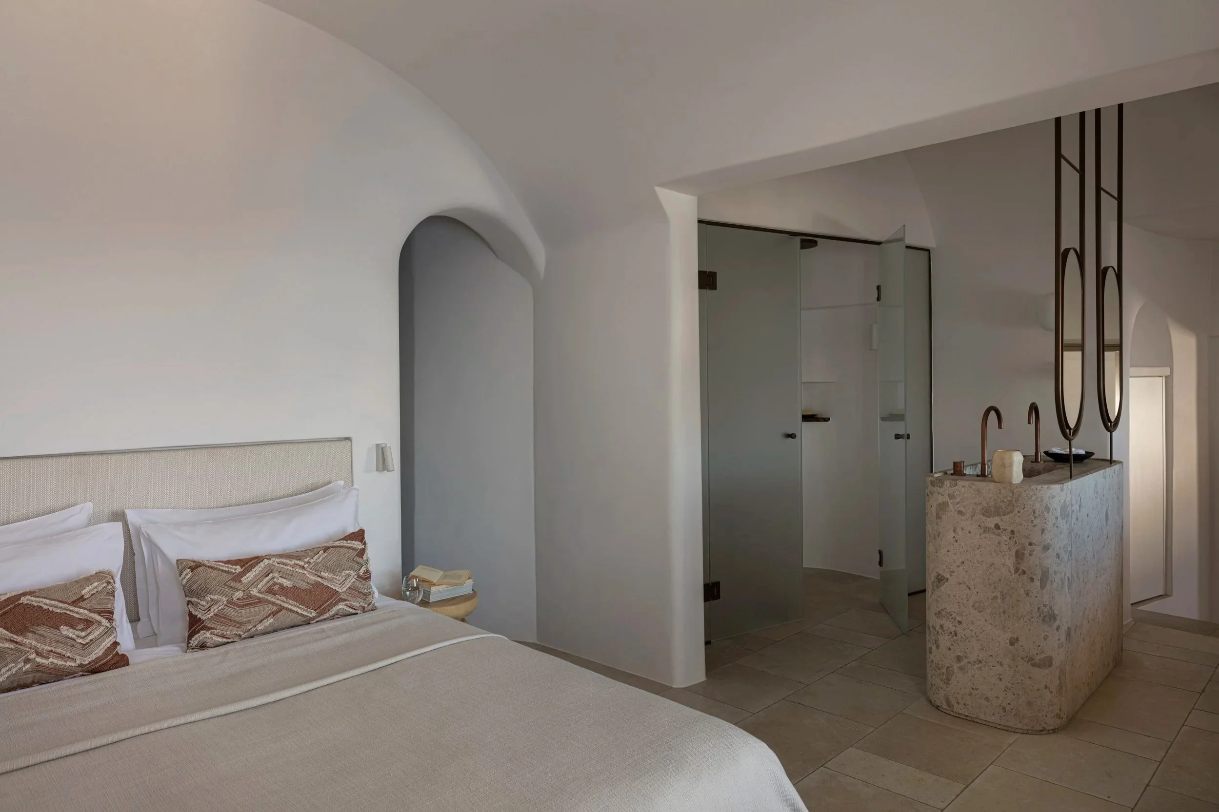 A bedroom with a bed featuring white pillows and beige bedding, a small nightstand with a book, and a stone sink with copper faucets in a minimalistic, modern design interior with neutral tones.