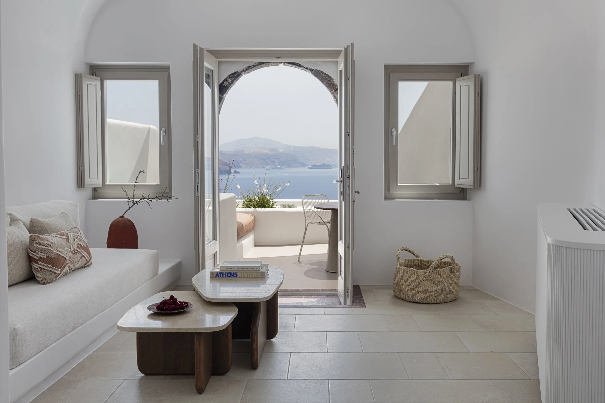 Interior of a bright white room with open glass doors leading to a balcony overlooking blue water and distant islands, with a small table and a sofa inside.