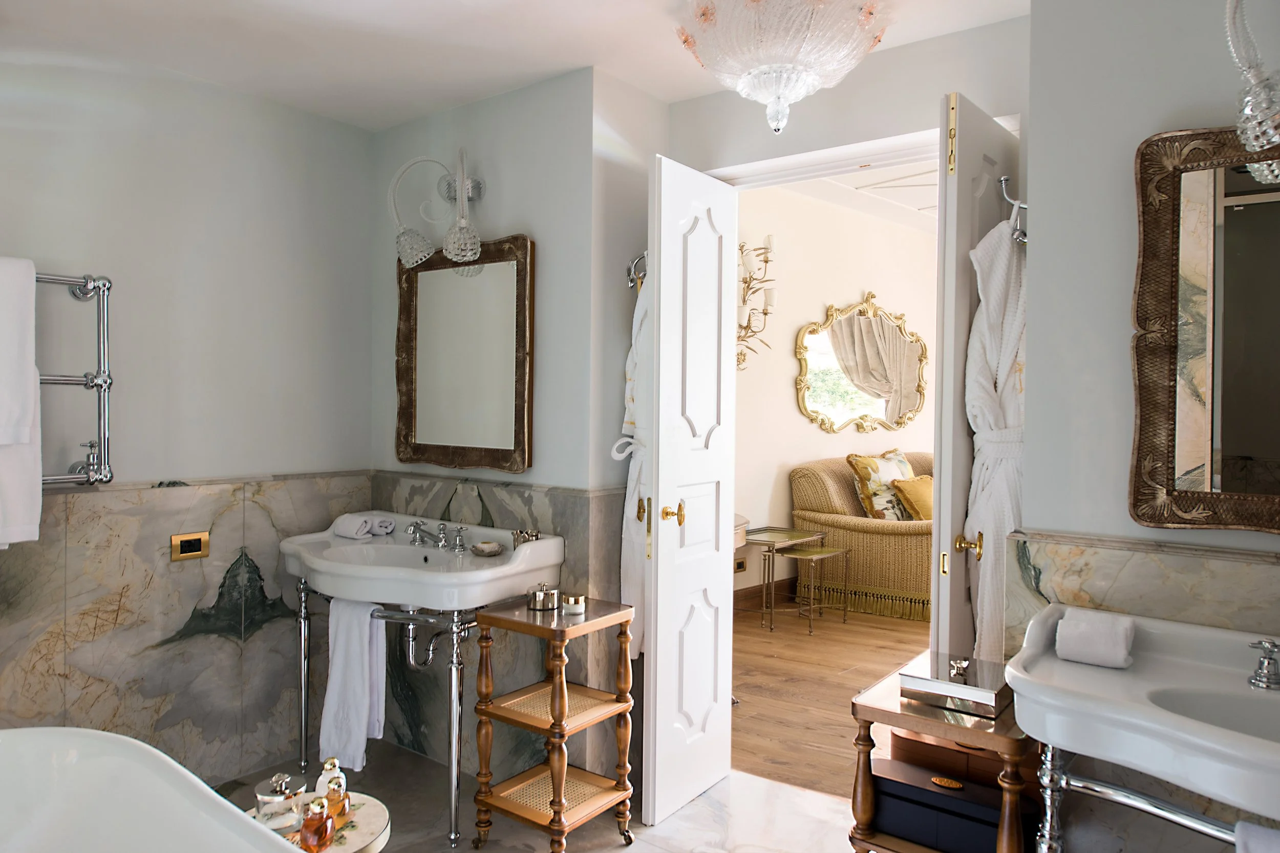 A bathroom with elegant decor, marble walls, two white sinks with mirrors above, and a view into a room with a gold-framed mirror and a sofa.