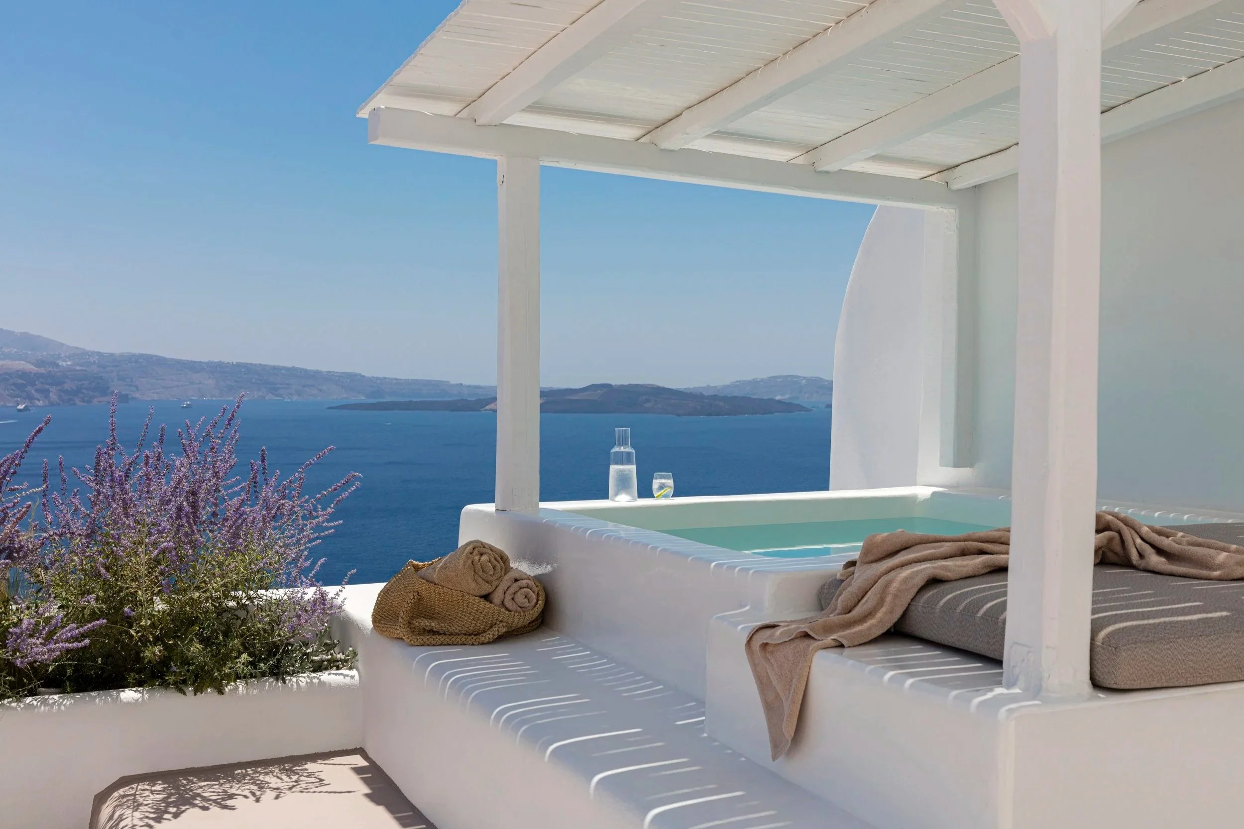 Greek-style outdoor terrace with a small pool, overlooking the sea, with towels and a glass of wine on a striped lounge chair, and purple flowers in the foreground.