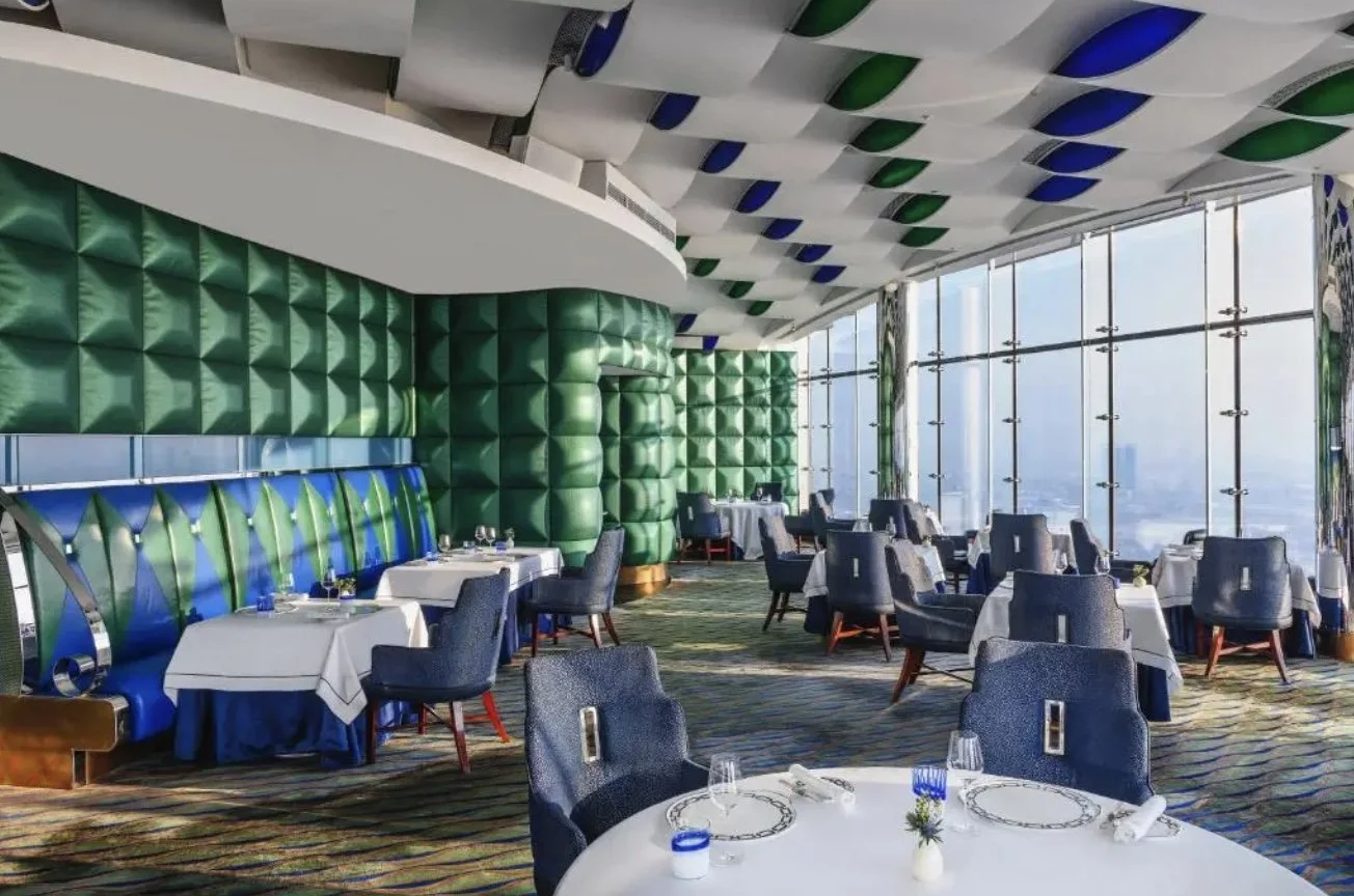 Interior of a modern restaurant with large glass windows, round tables with white tablecloths, blue and gray chairs, and colorful ceiling and wall decor in green, white, and blue.